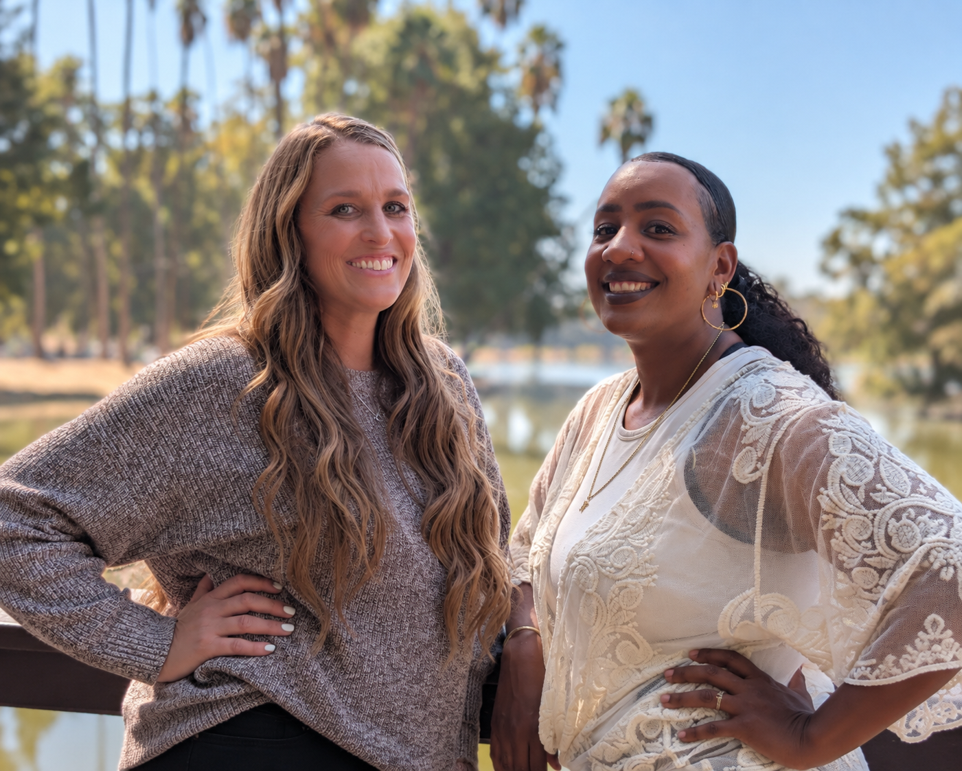 Two women smiling outdoors near a body of water with trees in the background, one with long wavy hair and the other with curly hair and earrings, wearing casual clothing.