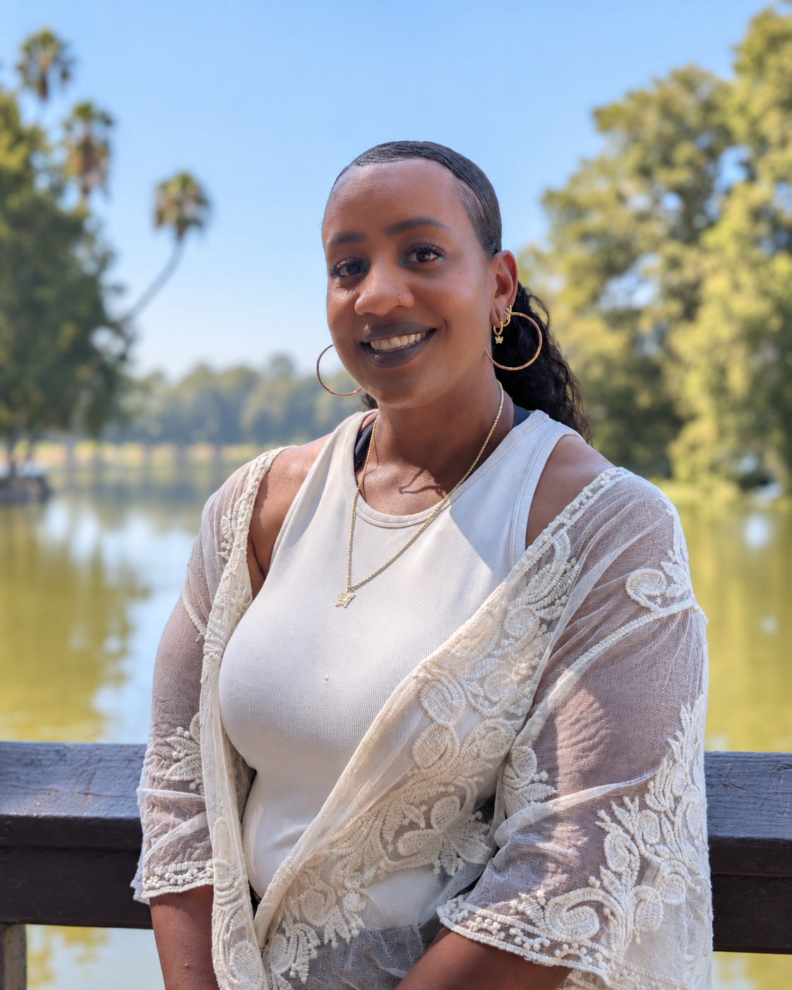 A woman with dark hair, wearing hoop earrings, a necklace, and a white lace-covered top, is smiling outdoors near a body of water with trees and a blue sky in the background.