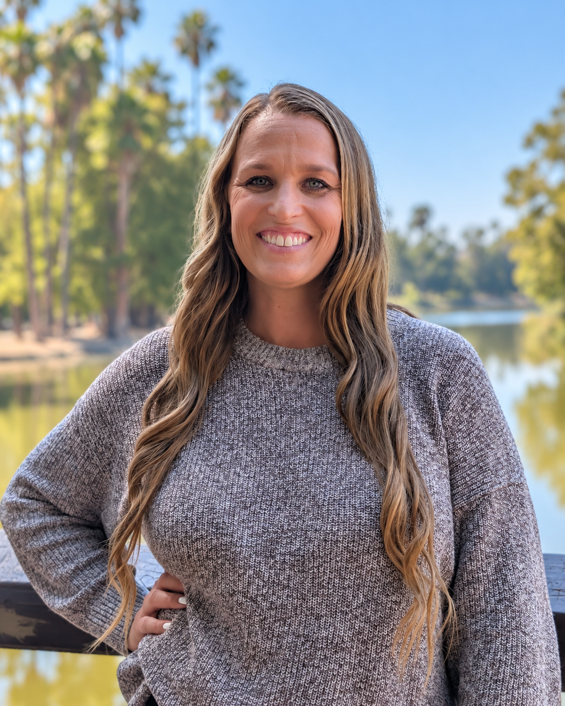 A woman with long wavy blonde hair, smiling, wearing a gray sweater, standing outdoors near a body of water with trees and clear blue sky in the background.