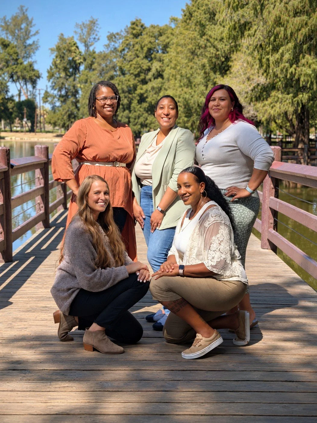 Six women standing and kneeling on a wooden bridge outdoors, smiling and posing for a group photo with trees and water in the background.