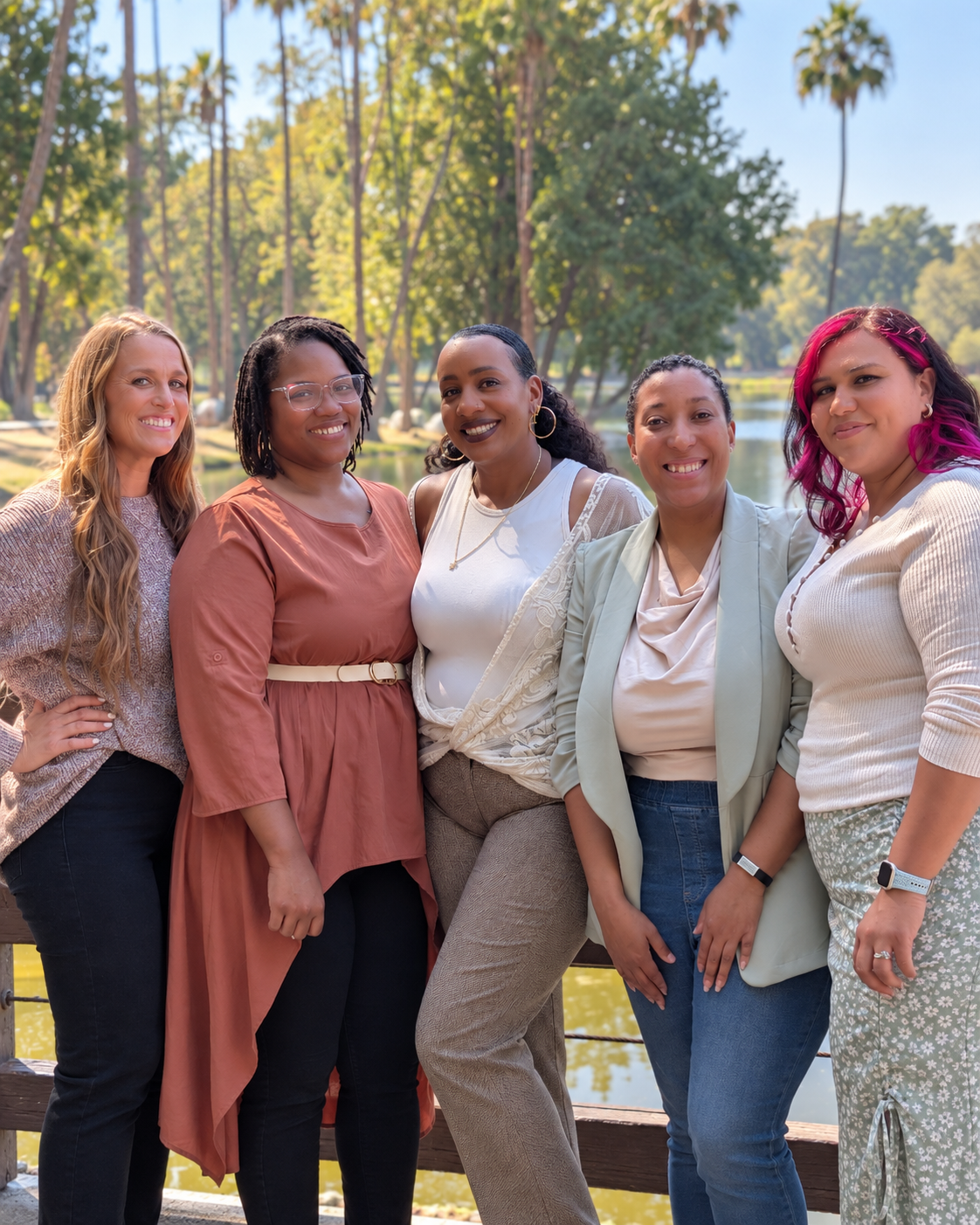 Group of five diverse women smiling together outdoors near a pond and trees.