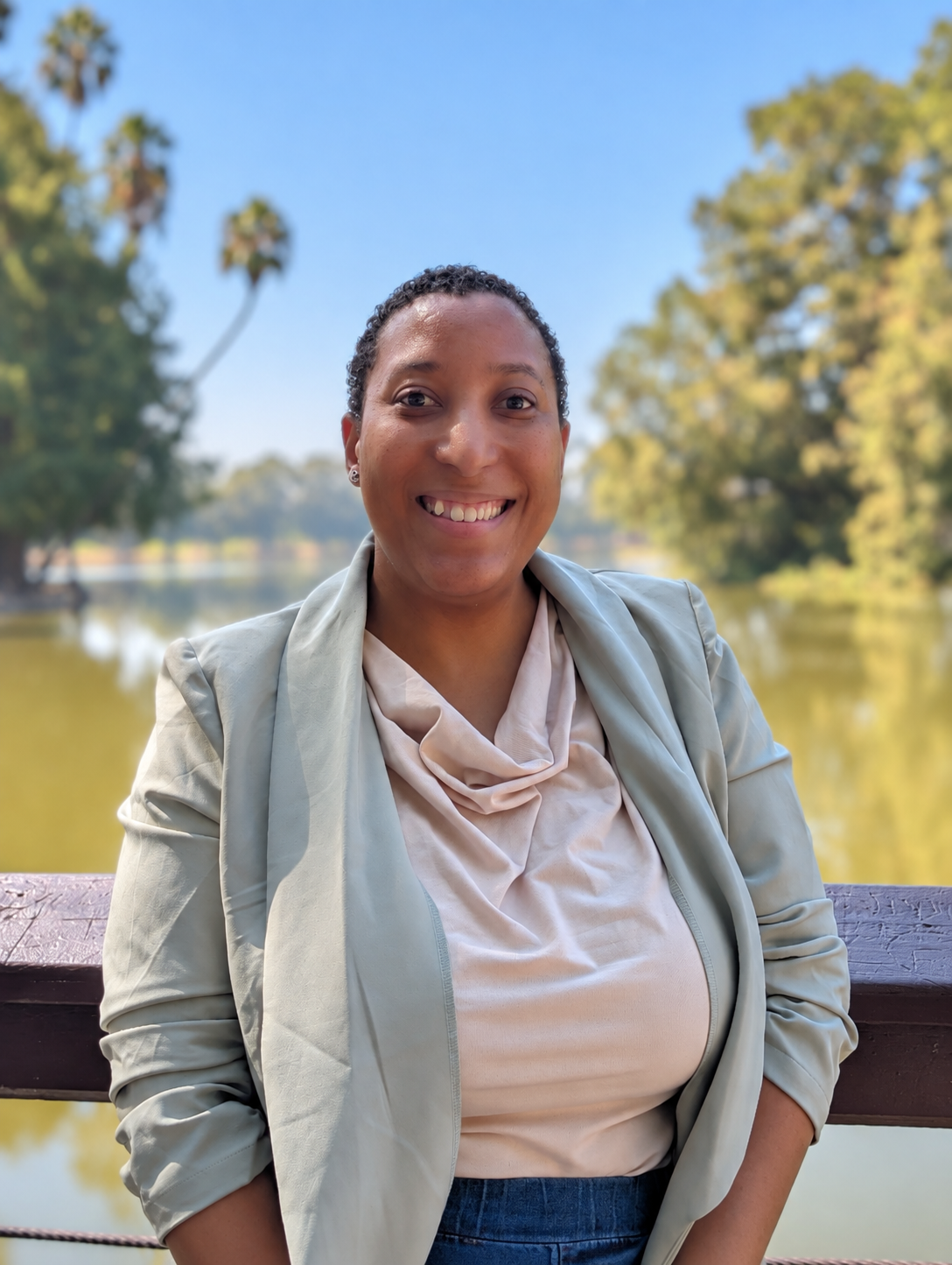 A woman smiling on a bridge by a lake, with trees and palm trees in the background.