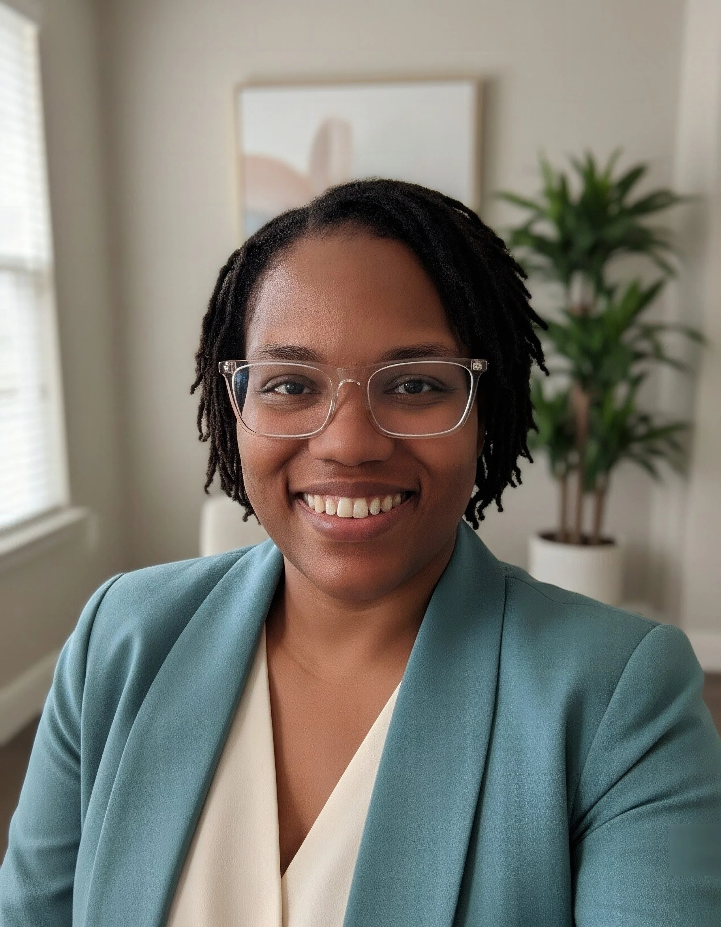 Close-up of a smiling African American woman with glasses, wearing a teal blazer over a white top, standing indoors with a blurred background of a potted plant and a framed picture.
