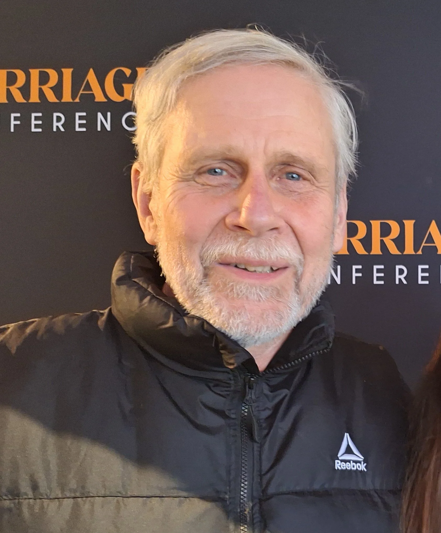 Tim Rovik, wearing a black Reebok jacket, standing in front of a black background with orange and white text at a conference.