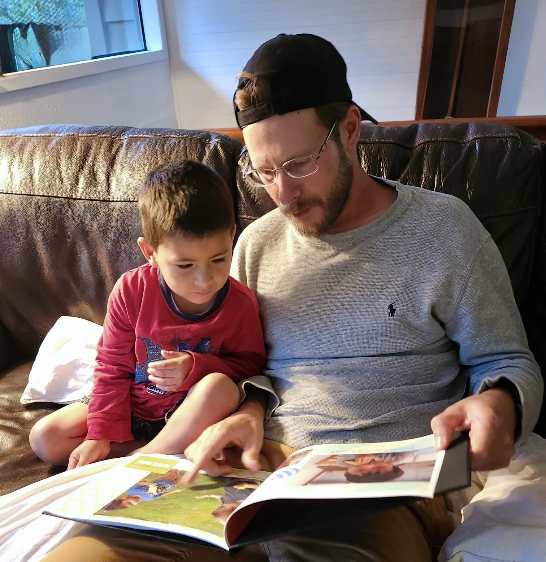 Dad and his son sit on a brown leather couch and are reading together.