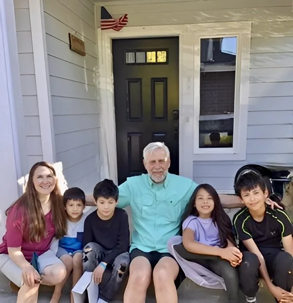 Tim Rovik and his wife sitting on a front porch of a house,  with their four multicultural grand children, smiling for the photo.