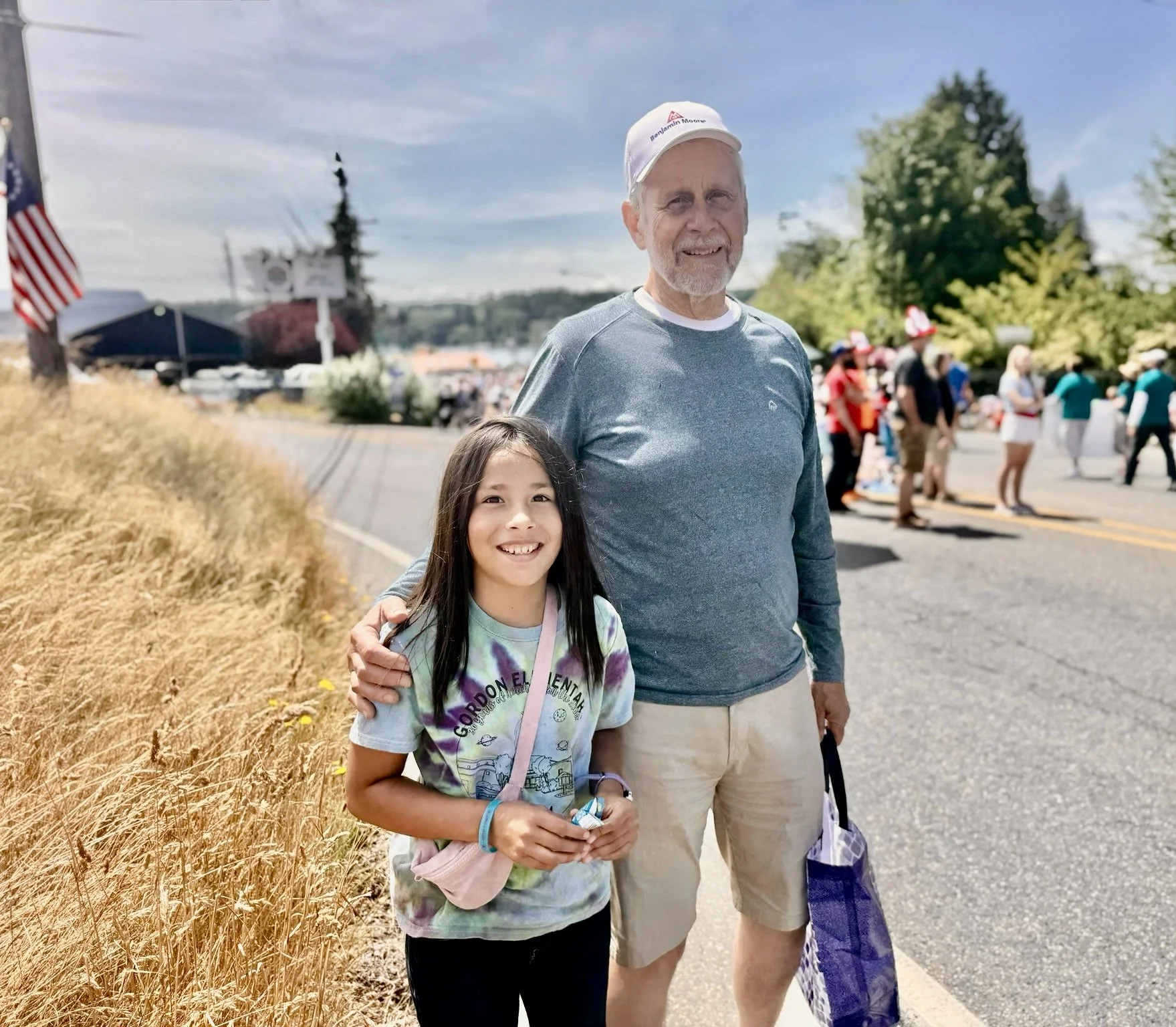 A smiling young granddaughter and Tim Rovik standing outdoors on a sunny day in Kingston WA with a crowd of people  in the background, near a street with a dry grassy area on the left.