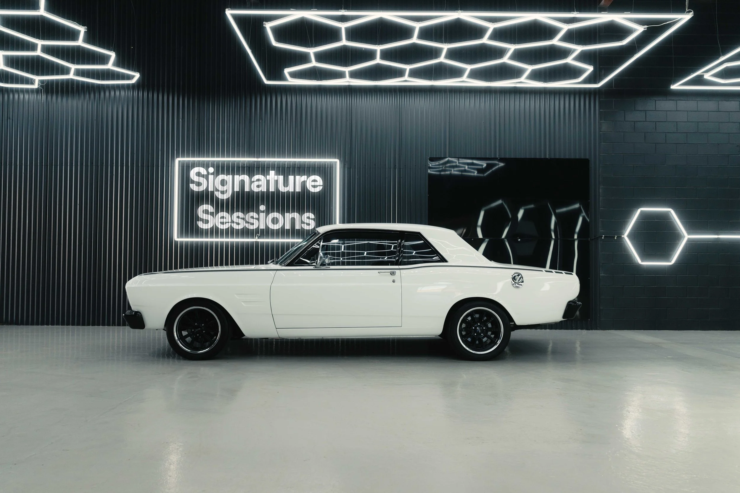A vintage white two-door coupe car with black wheels parked indoors against a dark wall with neon signage reading 'Signature Sessions' and geometric neon light decorations.