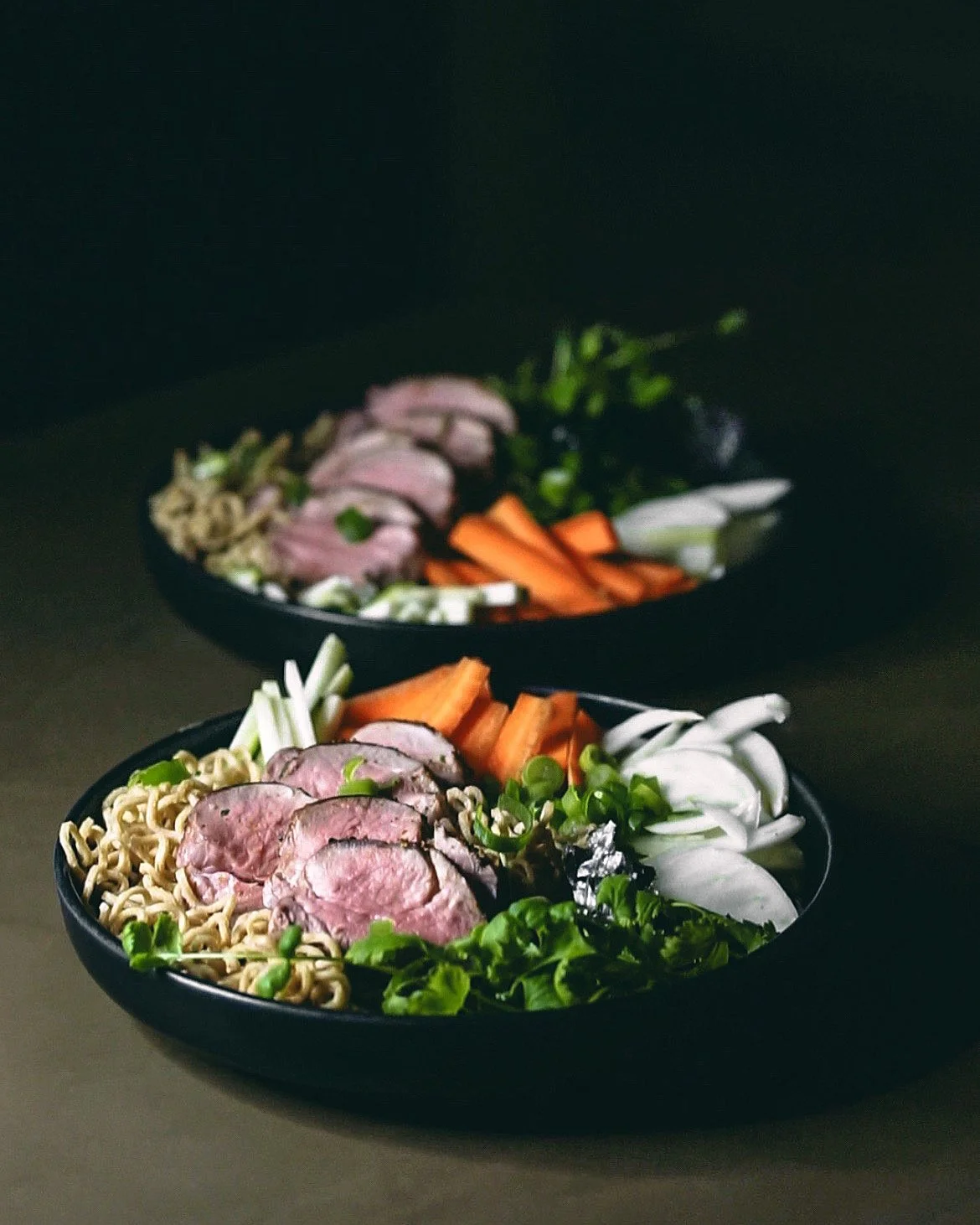 Two black bowls filled with ramen noodles, grilled meat slices, and fresh vegetables including green onions, carrots, and leafy greens on a dark surface.