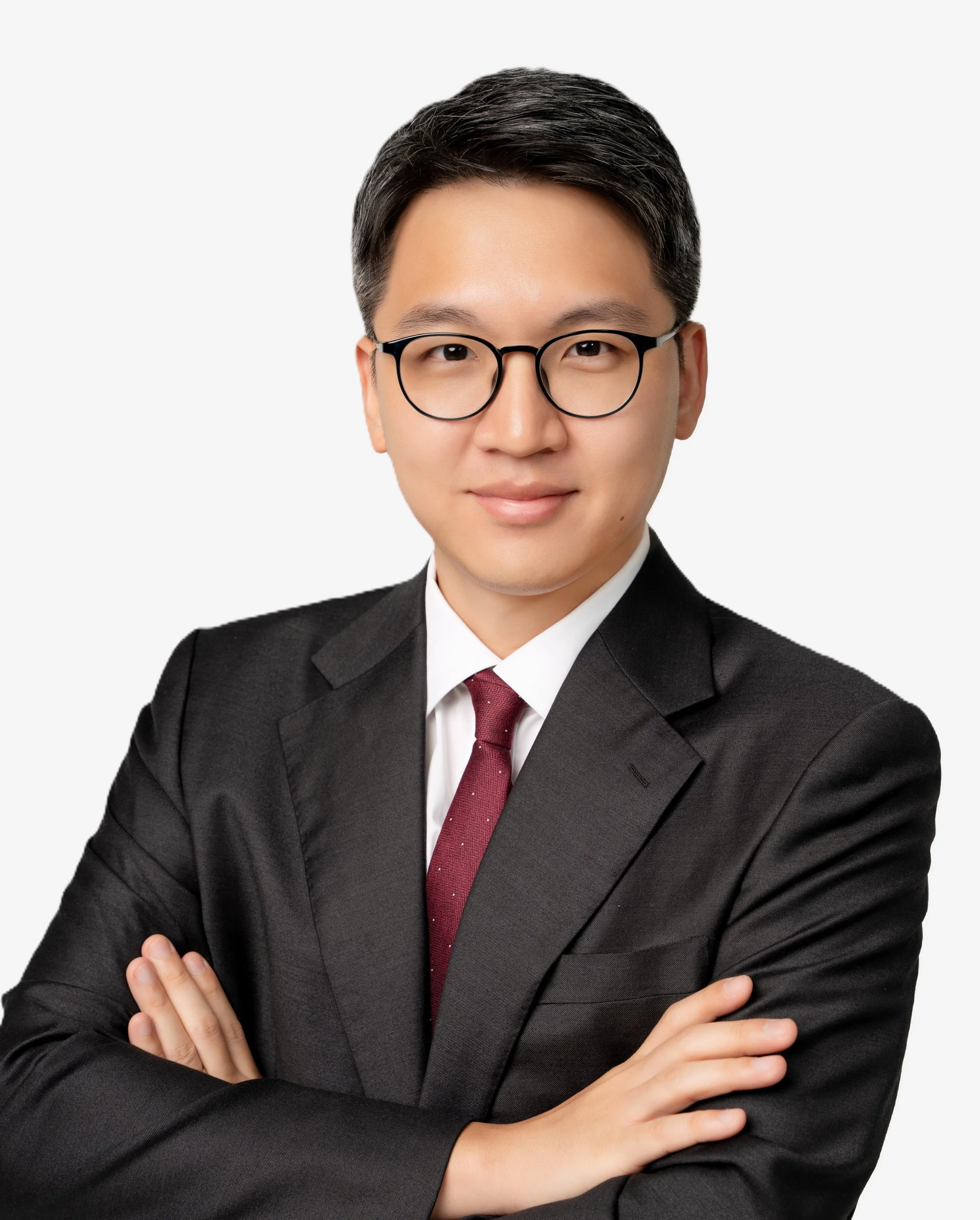 A young man with black hair, glasses, wearing a black suit, white dress shirt, and red tie, standing with arms crossed against a plain white background.