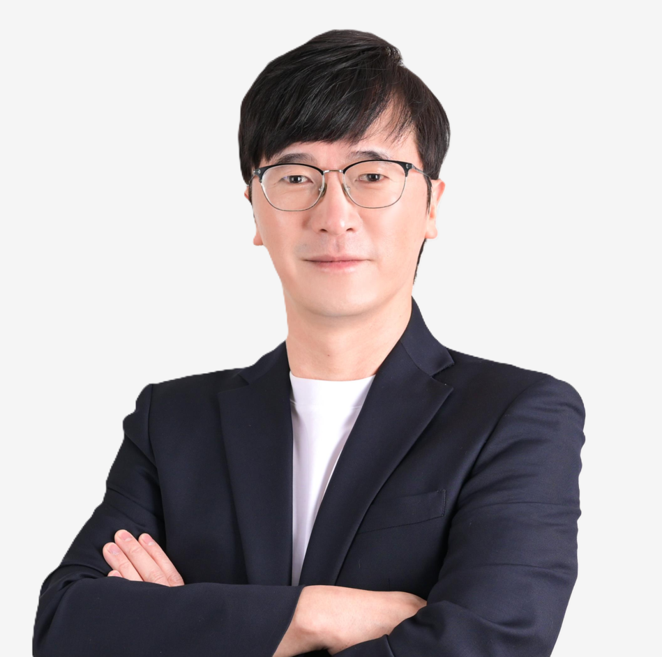 Portrait of a young man with black hair, glasses, wearing a black blazer and white shirt, crossing arms, smiling slightly, against a plain white background.