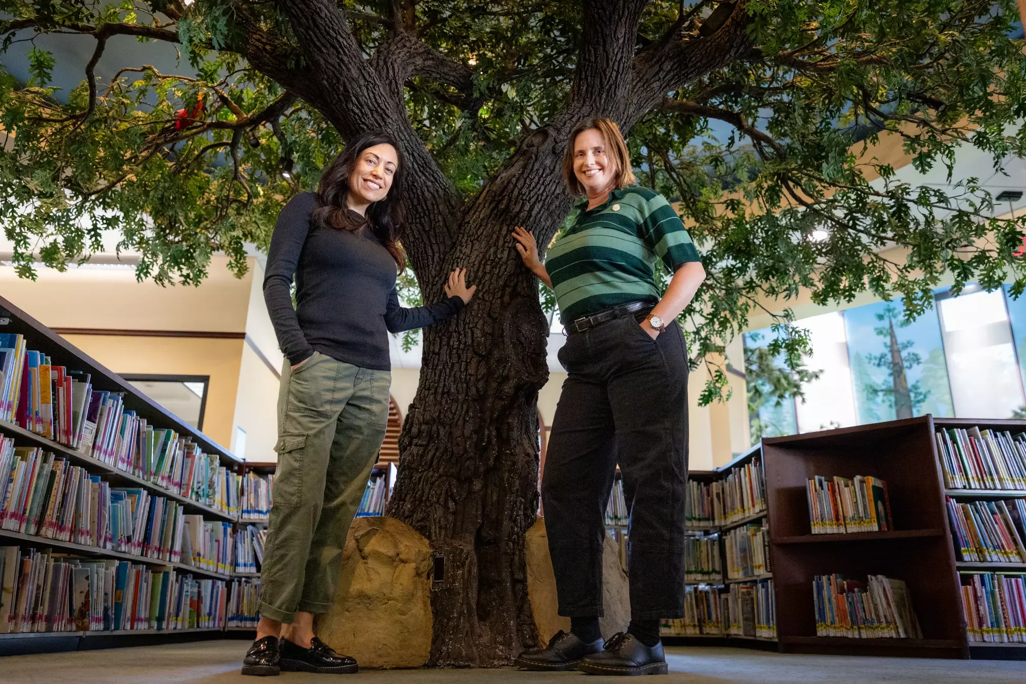 The two women who authored the book standing by a tree inside a library