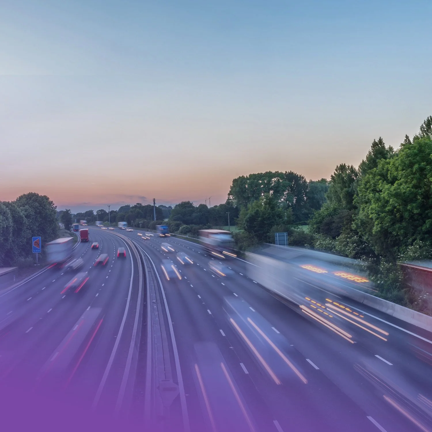 Long exposure photo of moving cars on a highway at dusk, with blurred lights and a backdrop of green trees and a colorful sky.