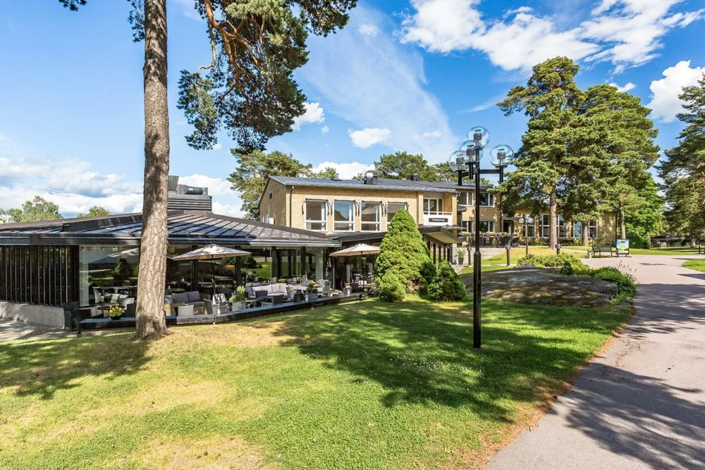 En modern byggnad med uteplats, omgivet av gröna träd och gräsyta under en klarblå himmel.