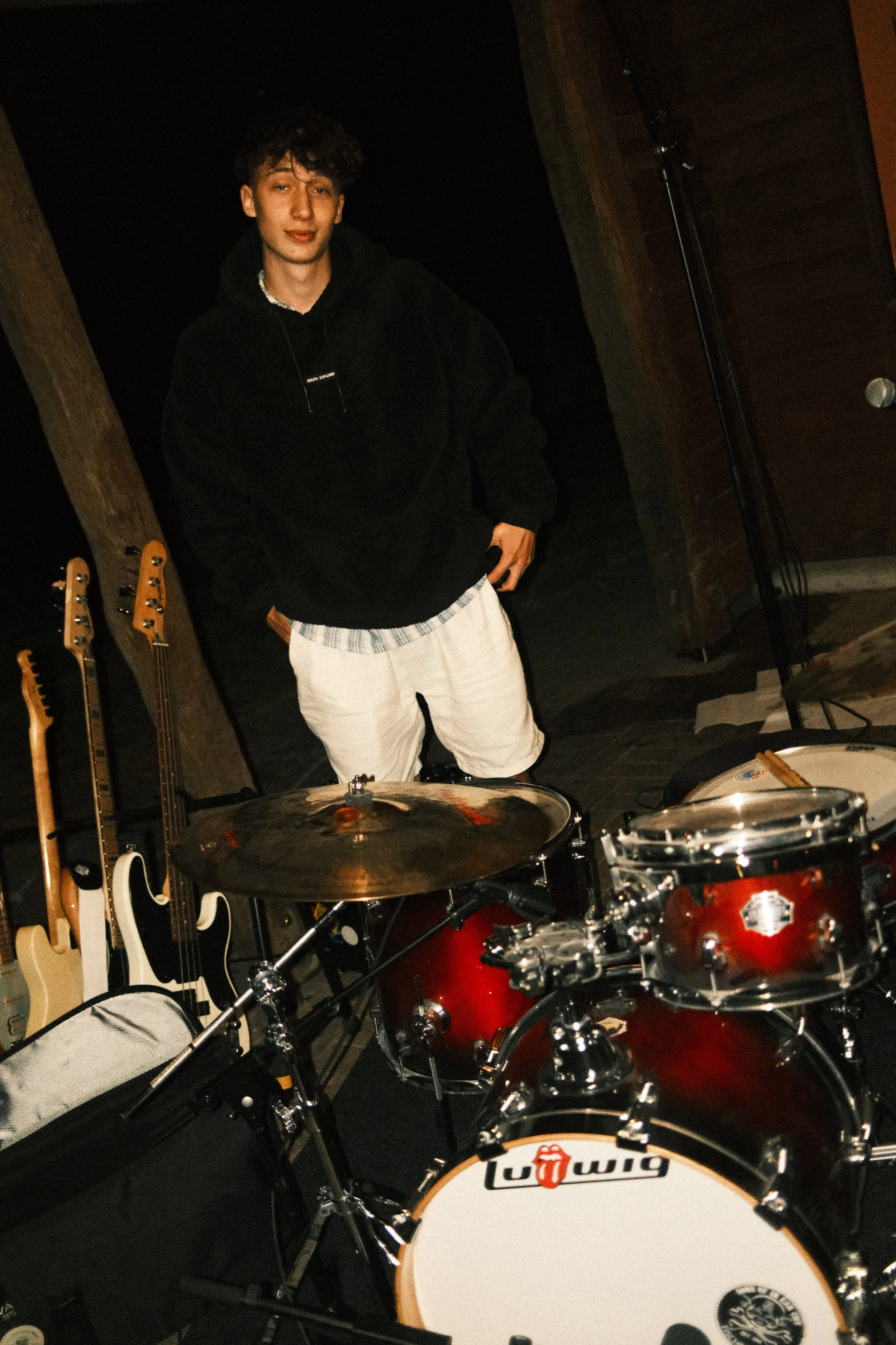 Junge Person steht hinter einem roten Schlagzeug in einem Musikstudio, umgeben von Gitarren, bei dunkler Beleuchtung.