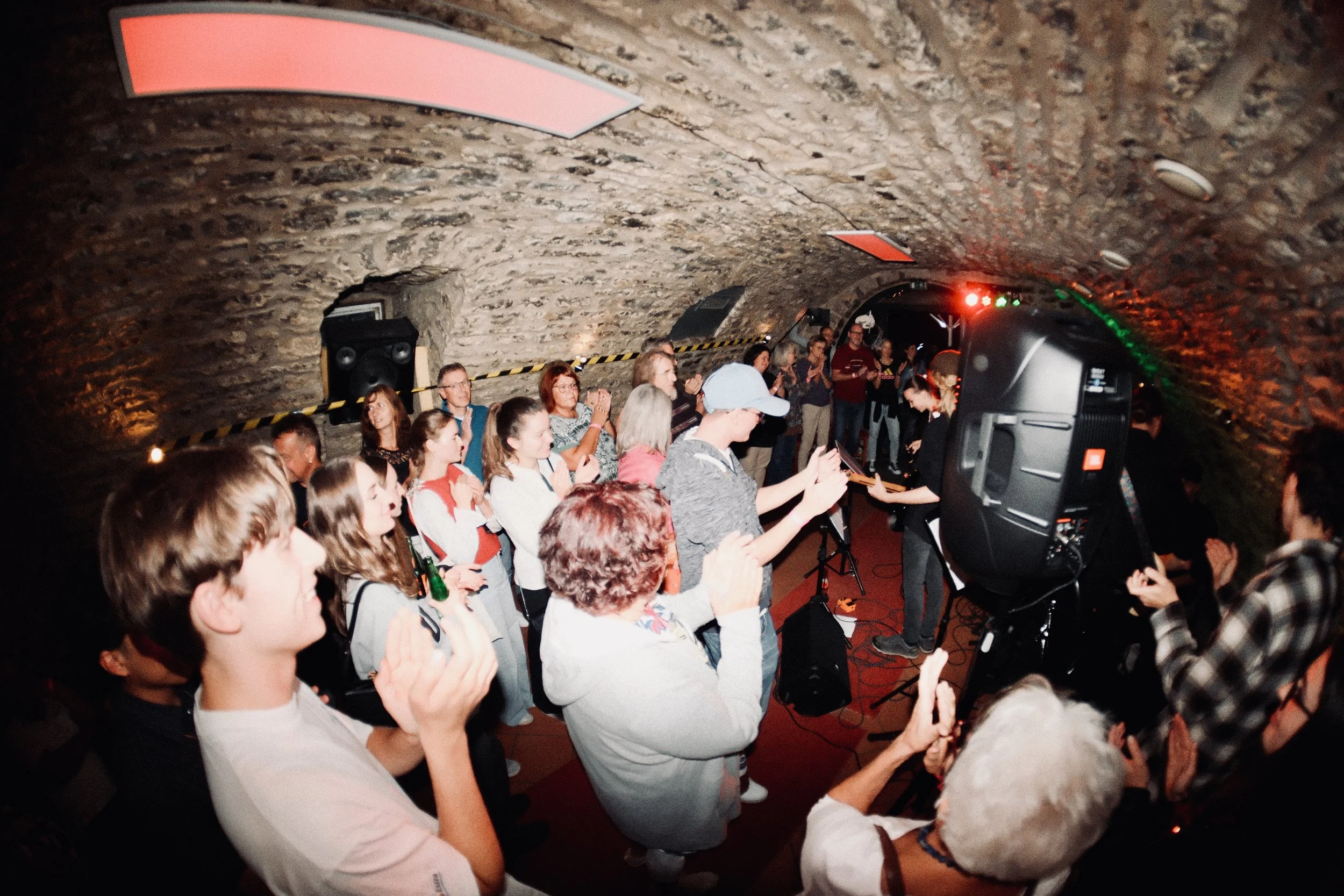 Menschen in einer Kellerkneipe mit Steinmauer, einige klatschen, andere filmen oder fotografieren, die Band optical flow aus Heilbronn spielt, bunte Lichter, Publikum steht eng zusammen und tanzt.