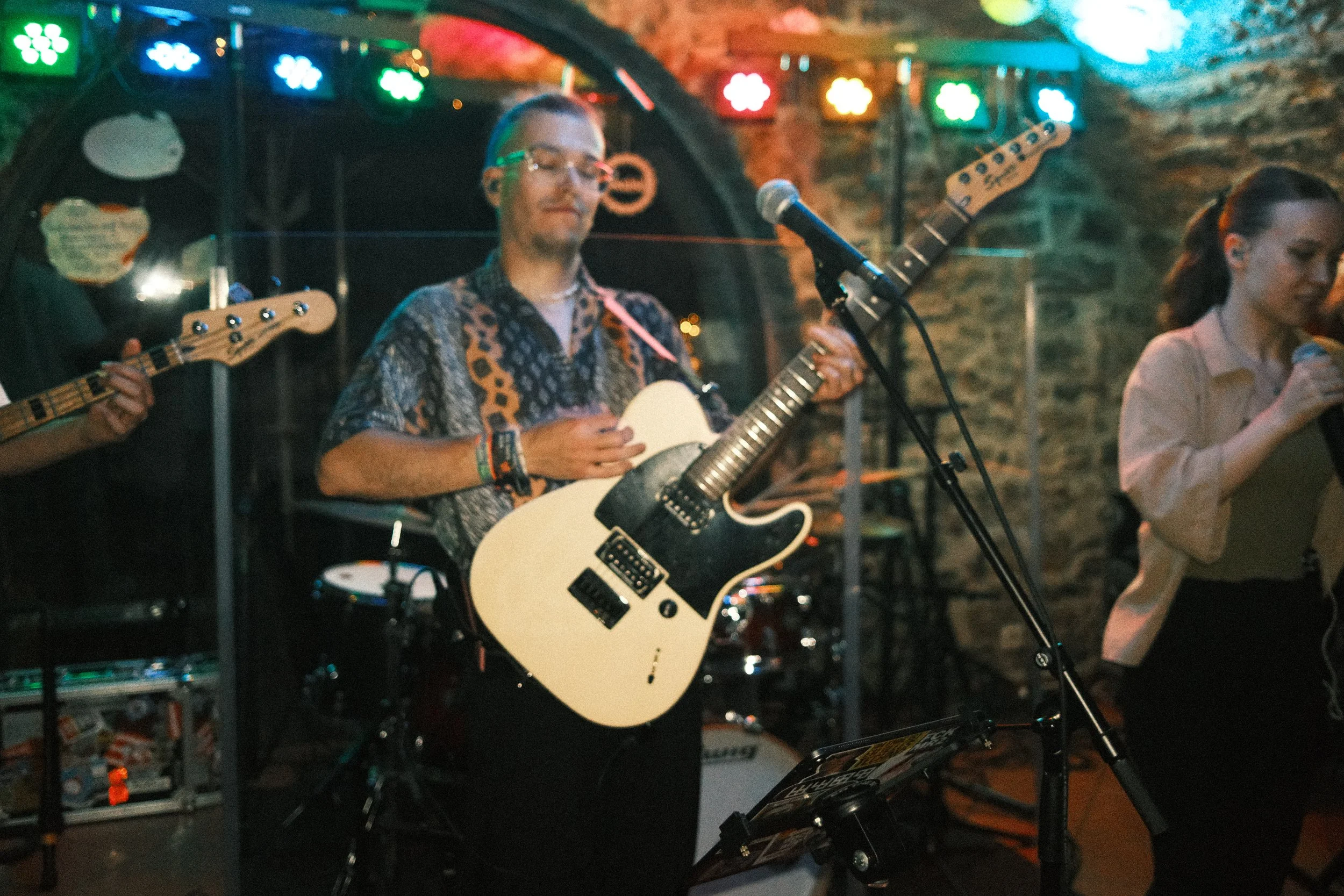 Musiker der Band optical flow spielen auf einer Bühne in einer Kneipe mit buntem Lichterhintergrund, einer hält eine elektrische Gitarre, eine andere singt in ein Mikrofon.