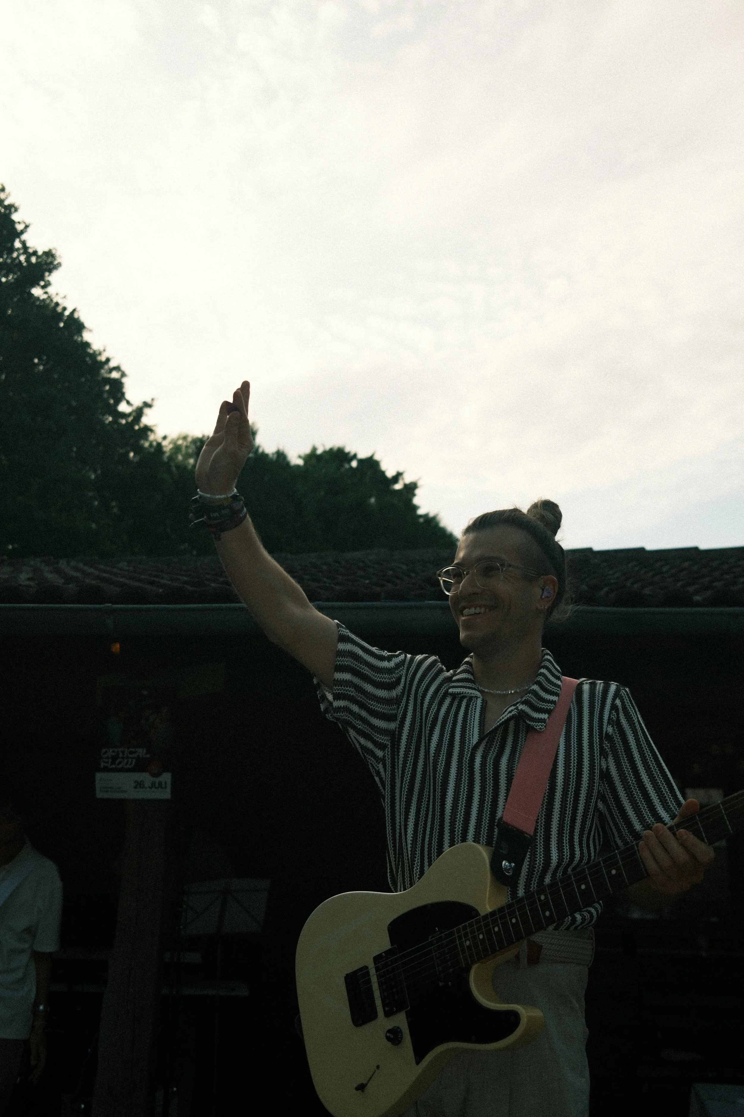 Junge Person mit Brille und geschlungener Haarfrisur spielt Gitarre, hebt die Hand und lächelt bei Sonnenuntergang, im Hintergrund Bäume.