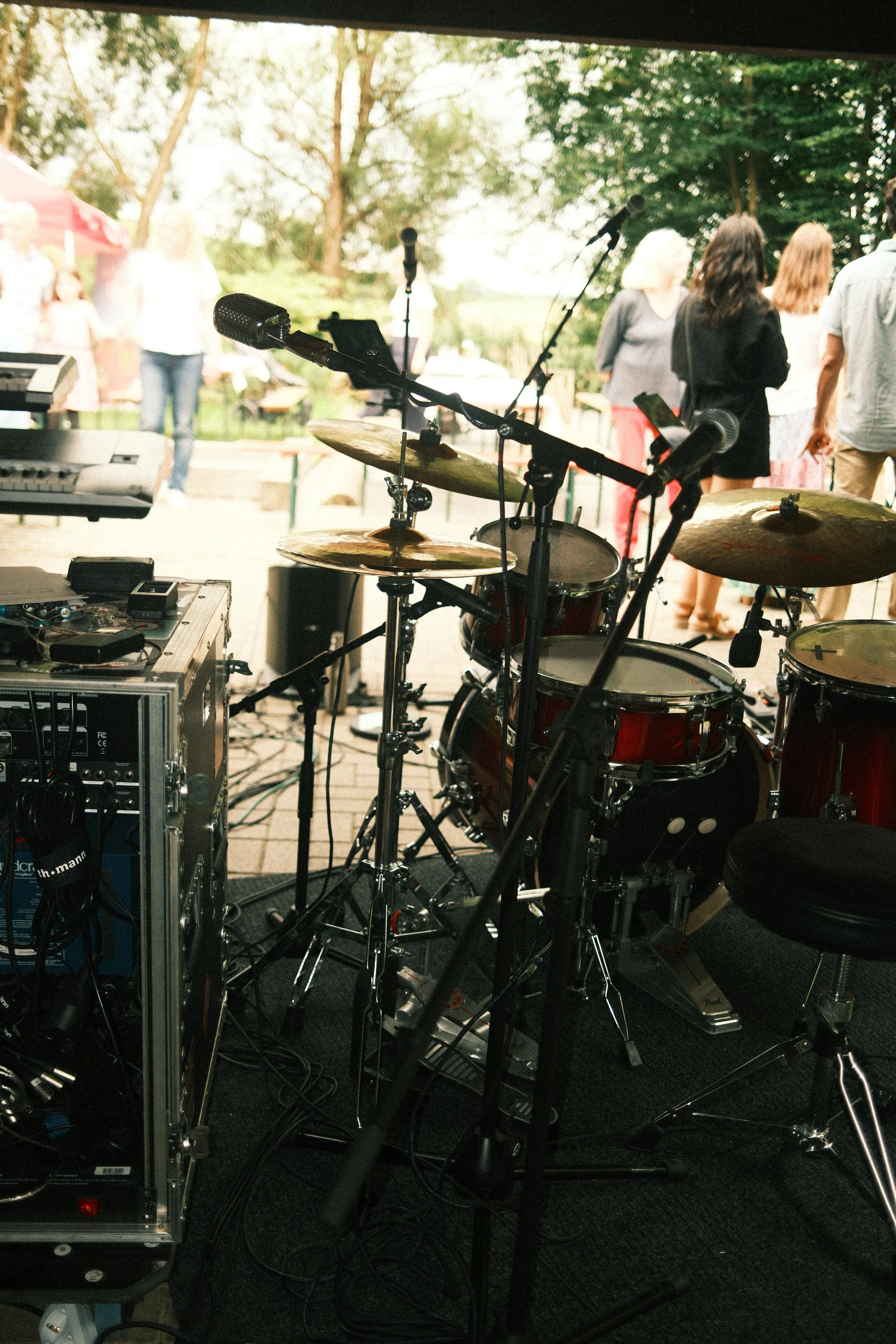 Blick auf eine Musikanlage mit Schlagzeug, Mikrophonen und Kabeln, im Hintergrund eine Gruppe von Menschen bei einem outdoor Event unter Bäumen.