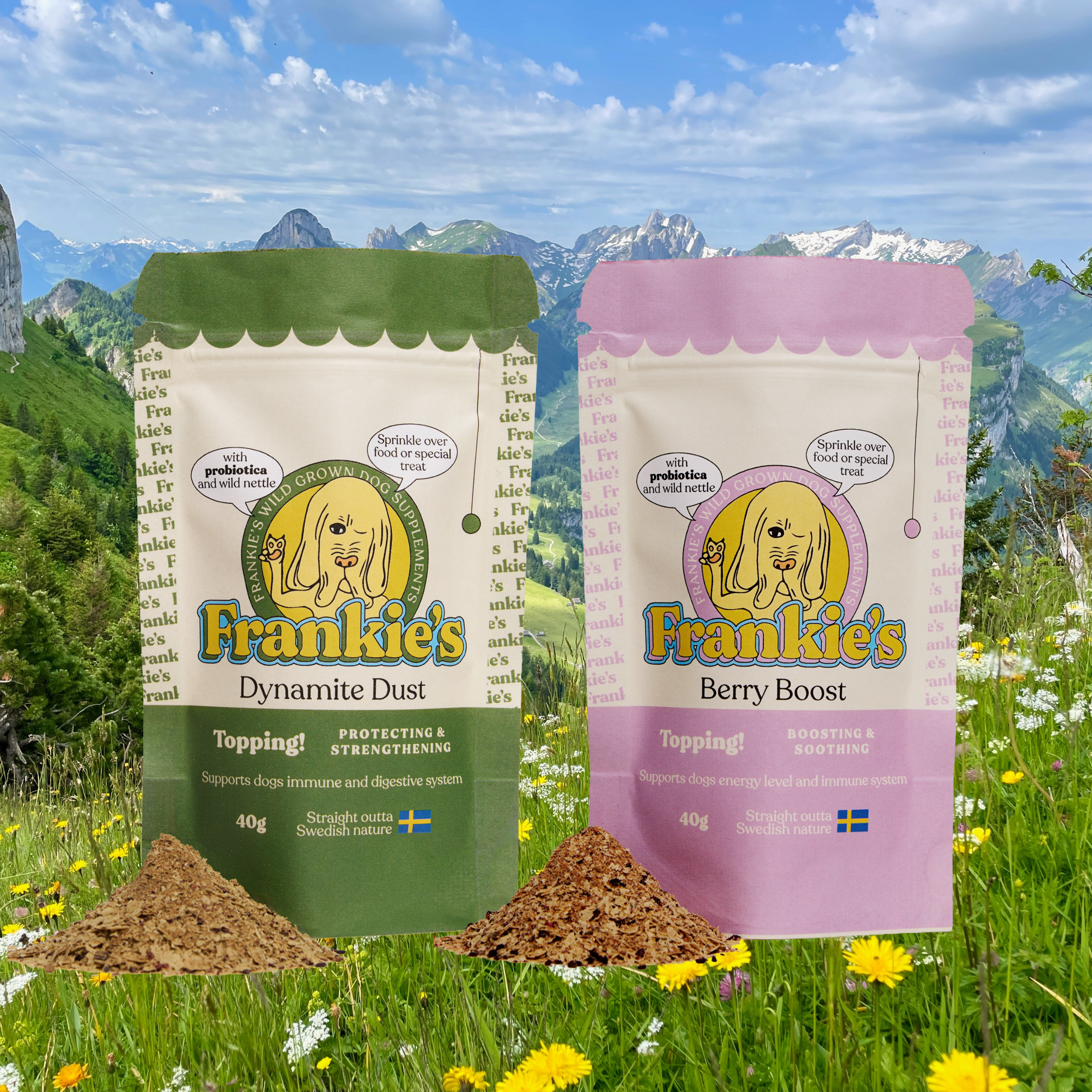 Two packets of Frankie's dog supplement powders, one labeled Dynamic Dust with green packaging and the other Berry Boost with pink packaging, placed behind small piles of powder in a lush green field with wildflowers, mountains, and a partly cloudy sky in the background.