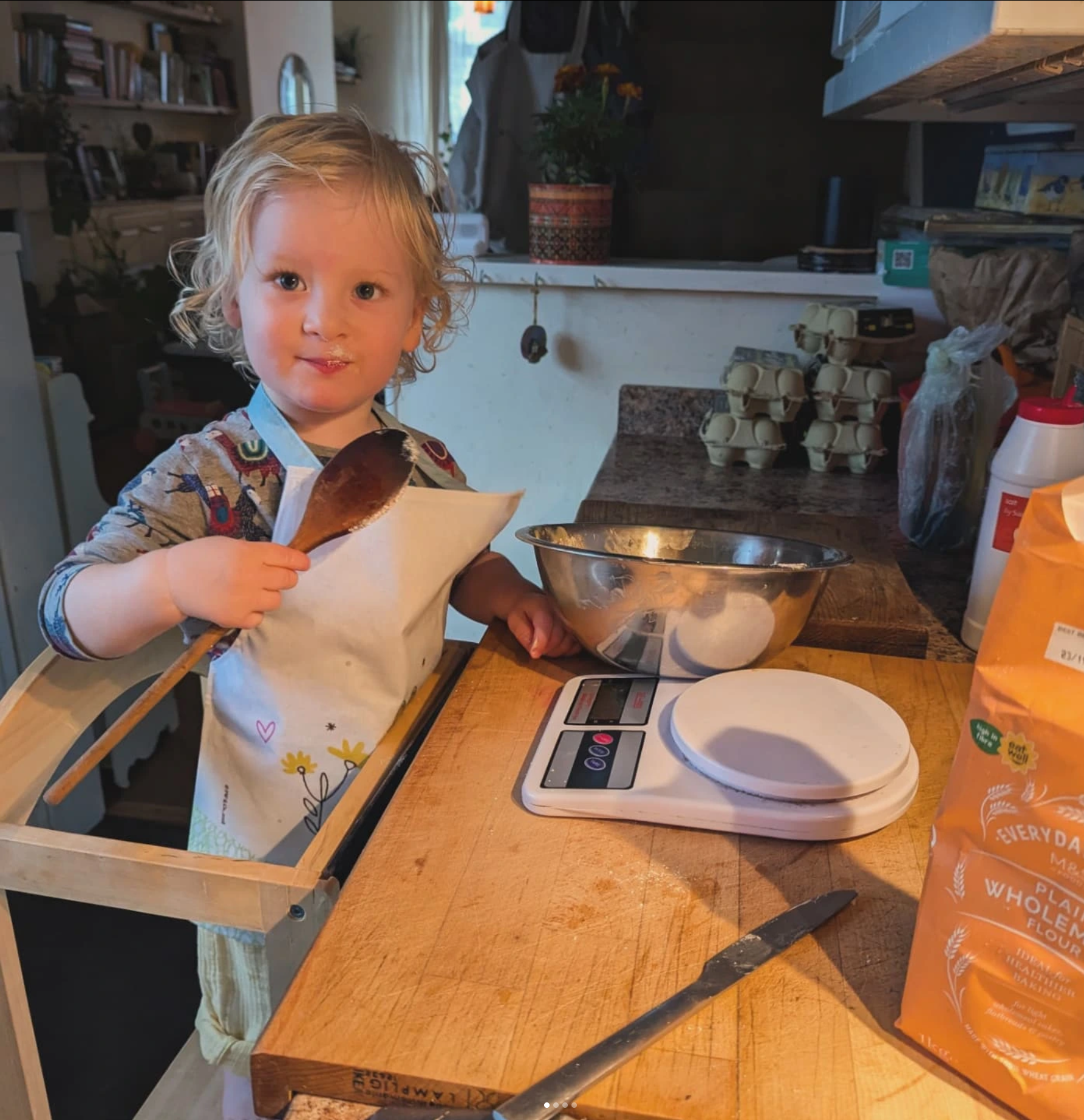 A young child with curly blonde hair wearing an apron, holding a wooden spoon, standing at a kitchen counter with a scale and a mixing bowl, preparing to bake.