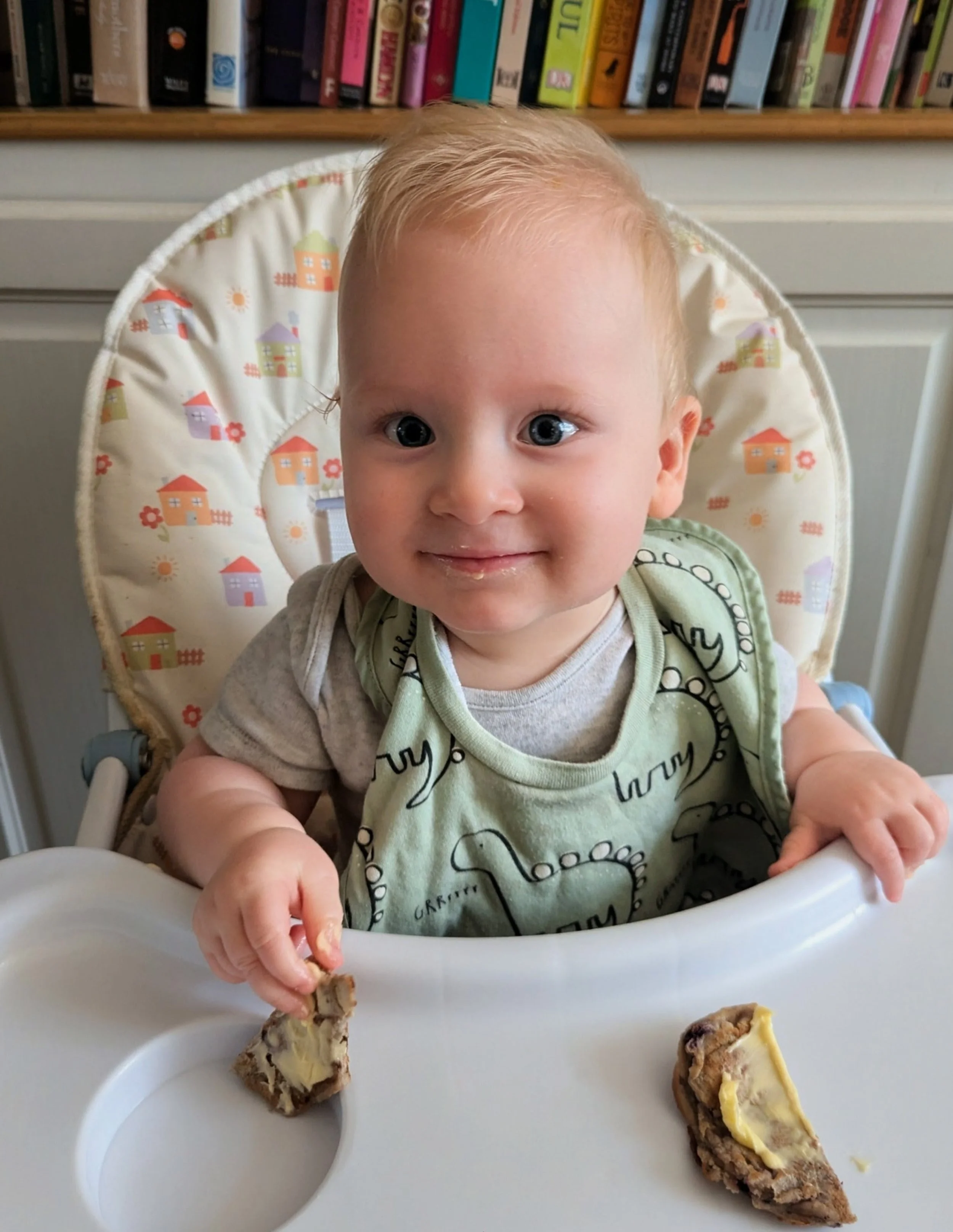 A smiling young child with blond hair and blue eyes sitting in a high chair at a table, holding a piece of food. The background shows a bookshelf with colorful books.