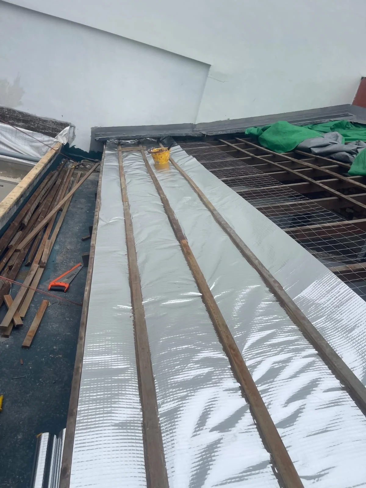 Construction site showing wooden beams, reflective insulation material, and roofing materials being installed on a building roof.