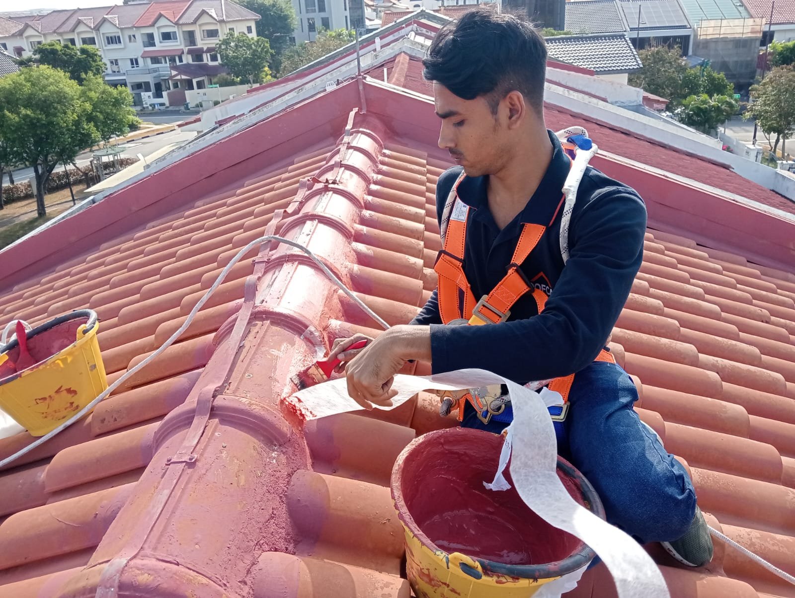 A man in work attire and safety harness works on painting a tiled roof with red paint, using a brush and containers of paint.