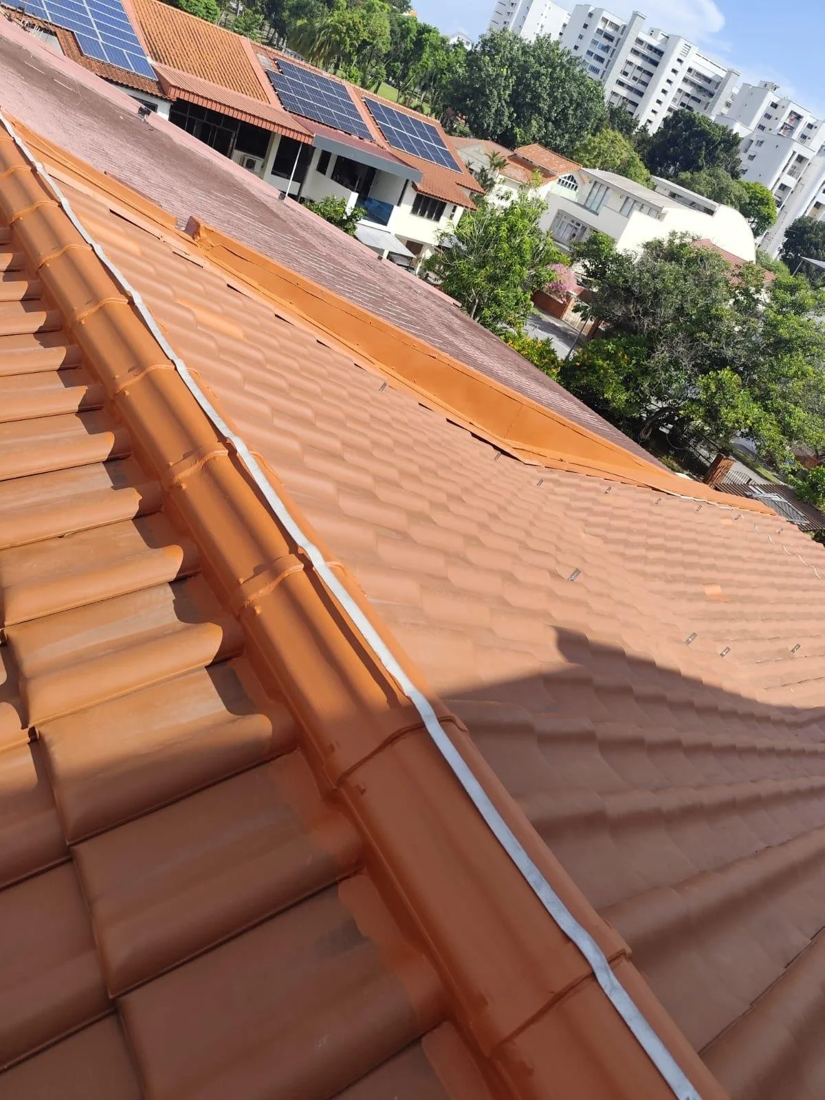 View of rooftops with solar panels, showing orange tiled roofs and a cityscape with high-rise buildings and green trees in the background.
