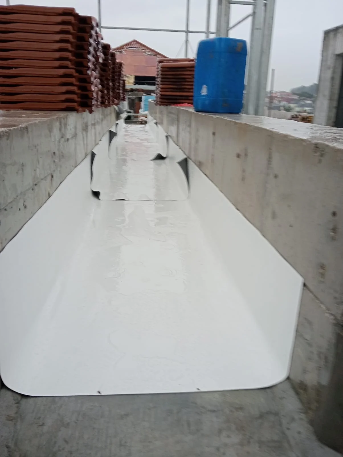 A close-up photo of a construction site showing a metal flashing installed between two concrete structures, with stacks of roof tiles and a blue water container in the background on a cloudy day.