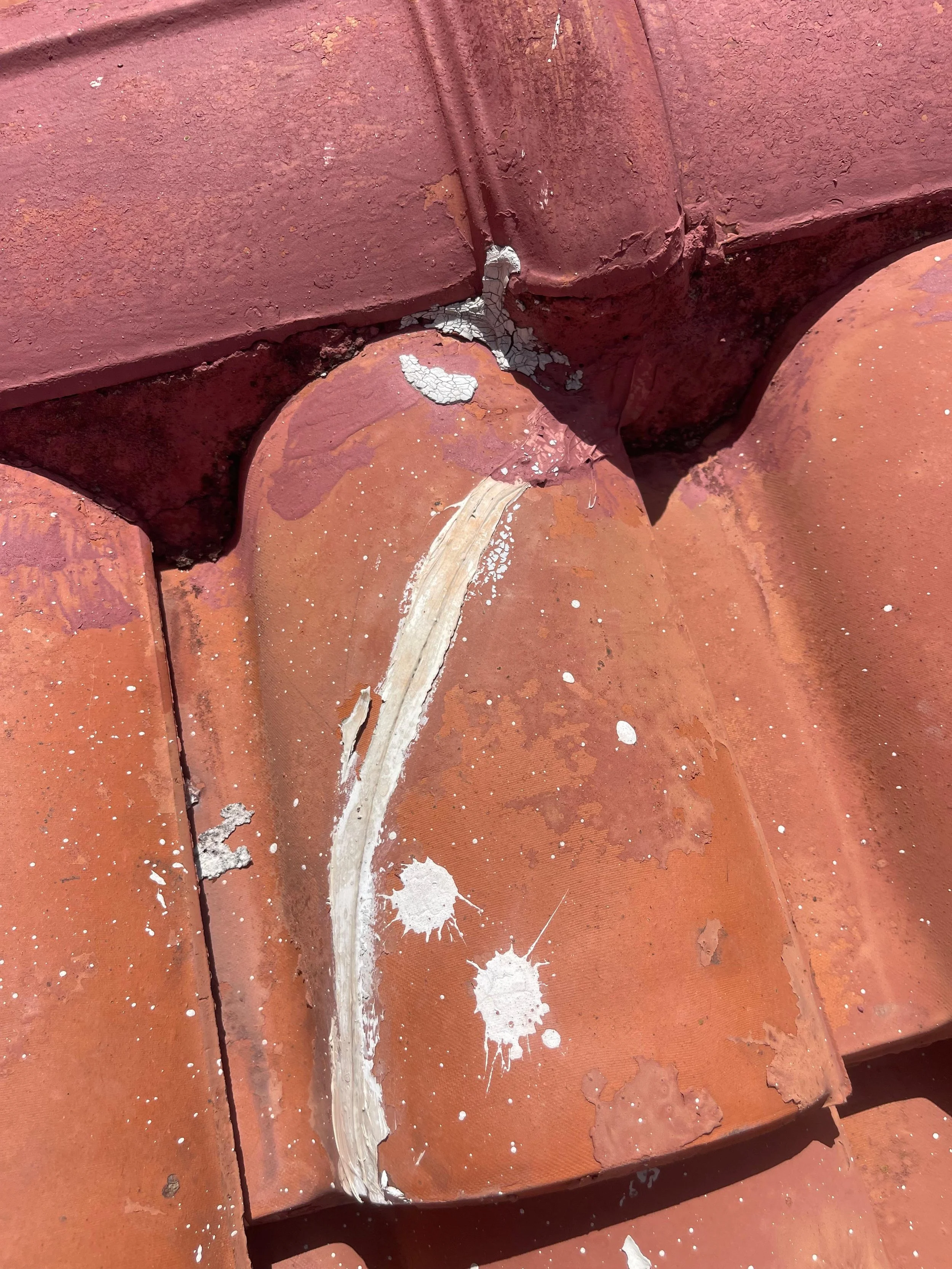 Close-up image of a damaged, weathered red clay roof tile with a crack running along its surface, paint splatters and dirt.