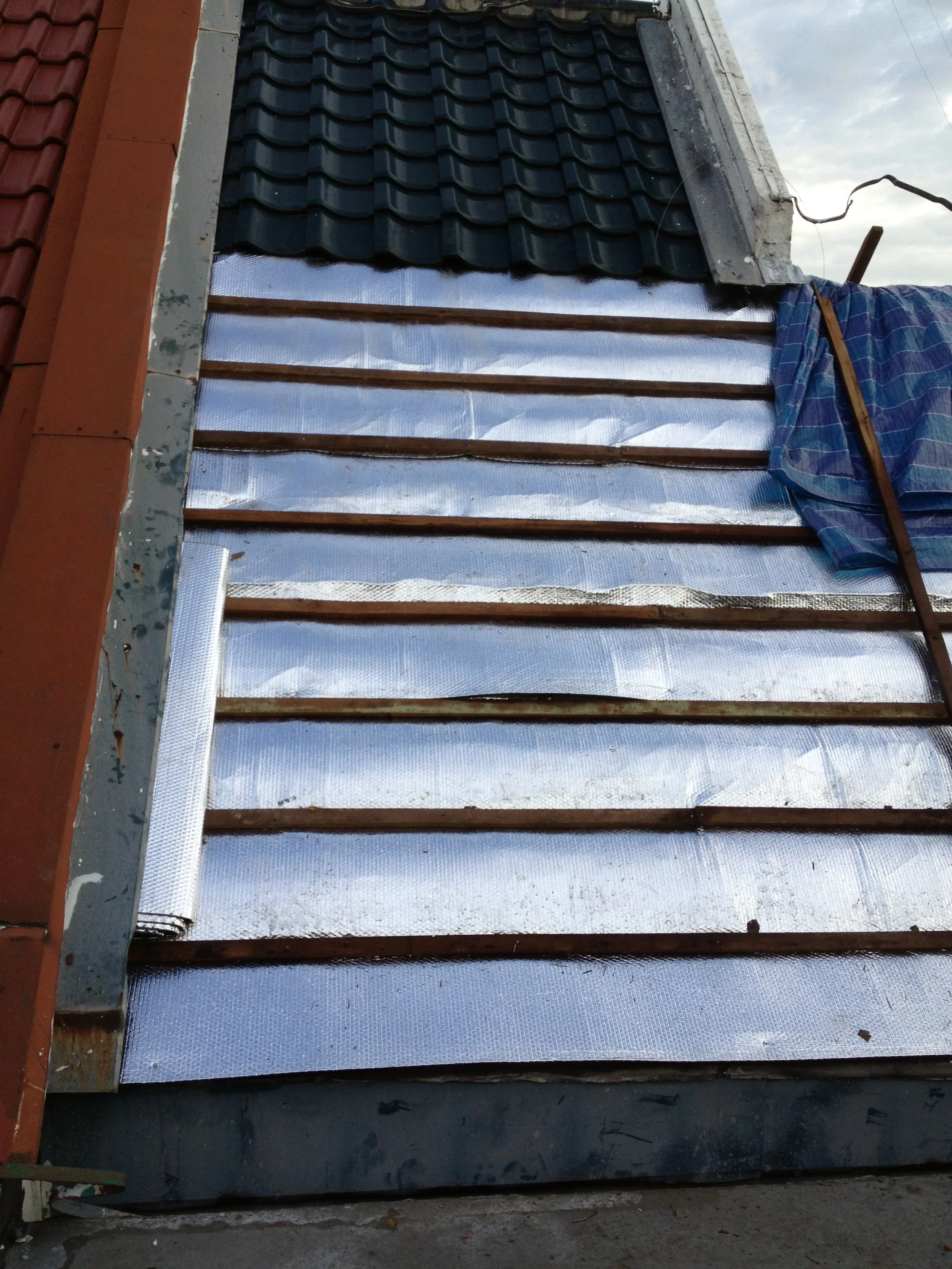 Construction of a building exterior with reflective insulation and ventilation gaps, partially covered by blue tarp and roofing tiles.