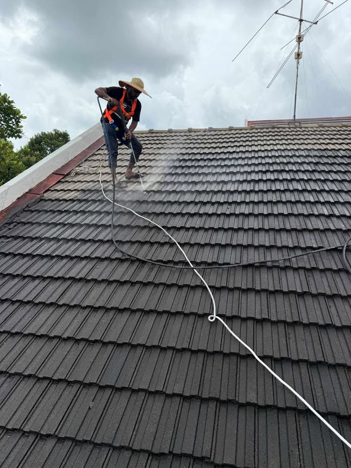A worker cleaning roof shingles with a power washer on a cloudy day, wearing a straw hat, safety harness, and dark clothing.