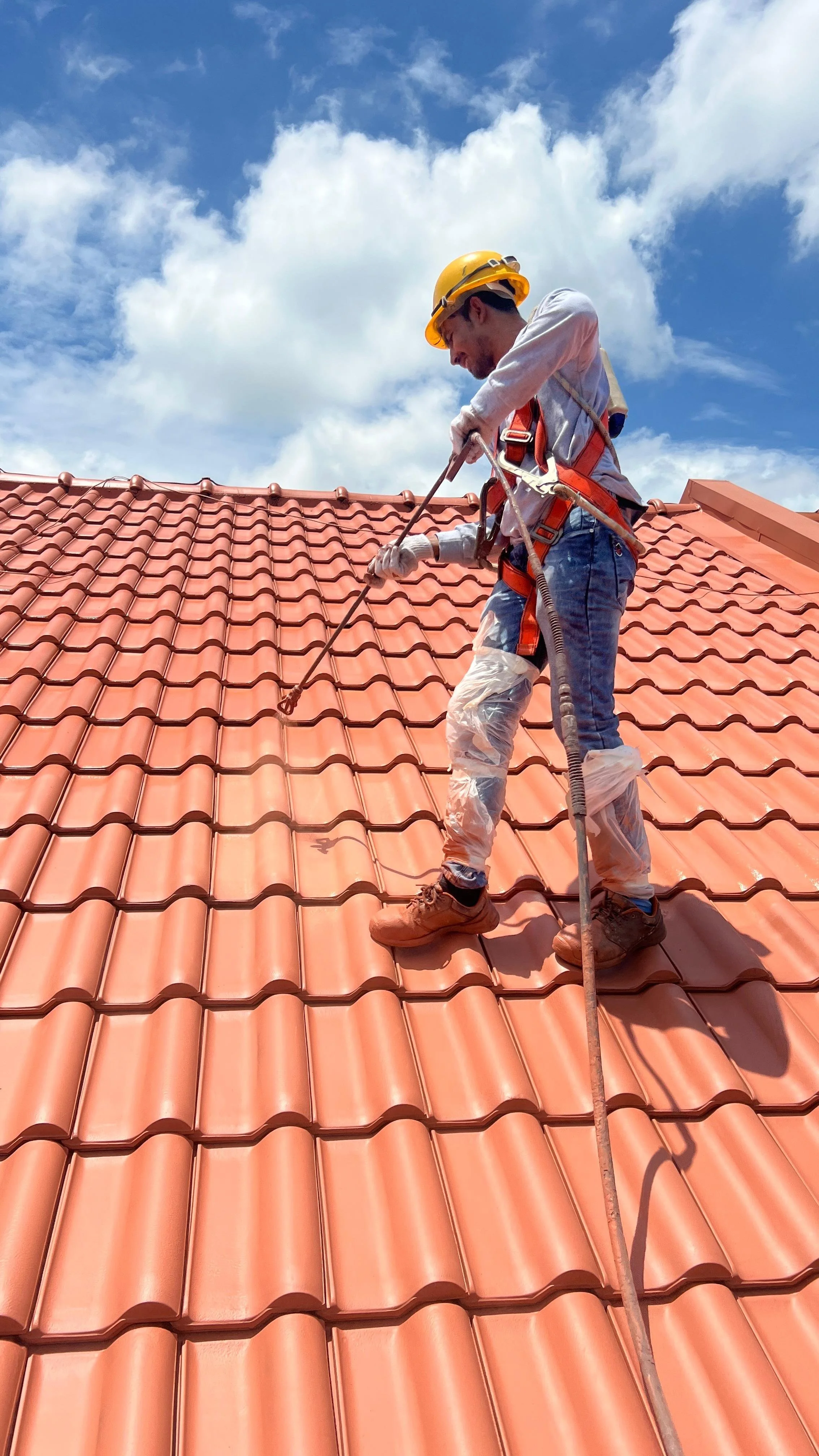 A construction worker wearing a yellow safety helmet, harness, and protective gear is standing on a red-tiled roof, using a power tool to work on the tiles under a partly cloudy sky.
