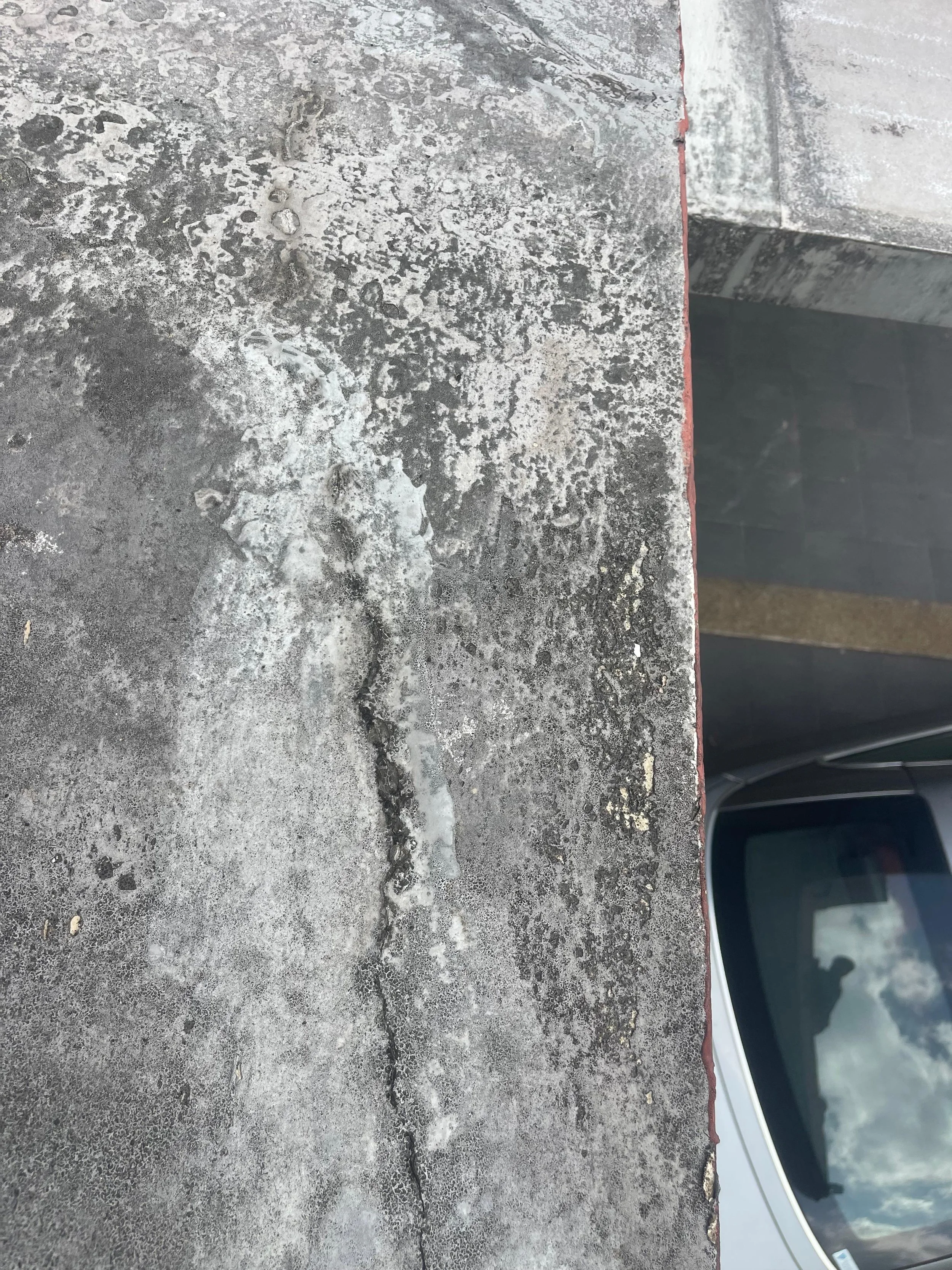 Close-up of a weathered concrete surface with cracks and patches, beside a metal edge with a car window visible in the background.