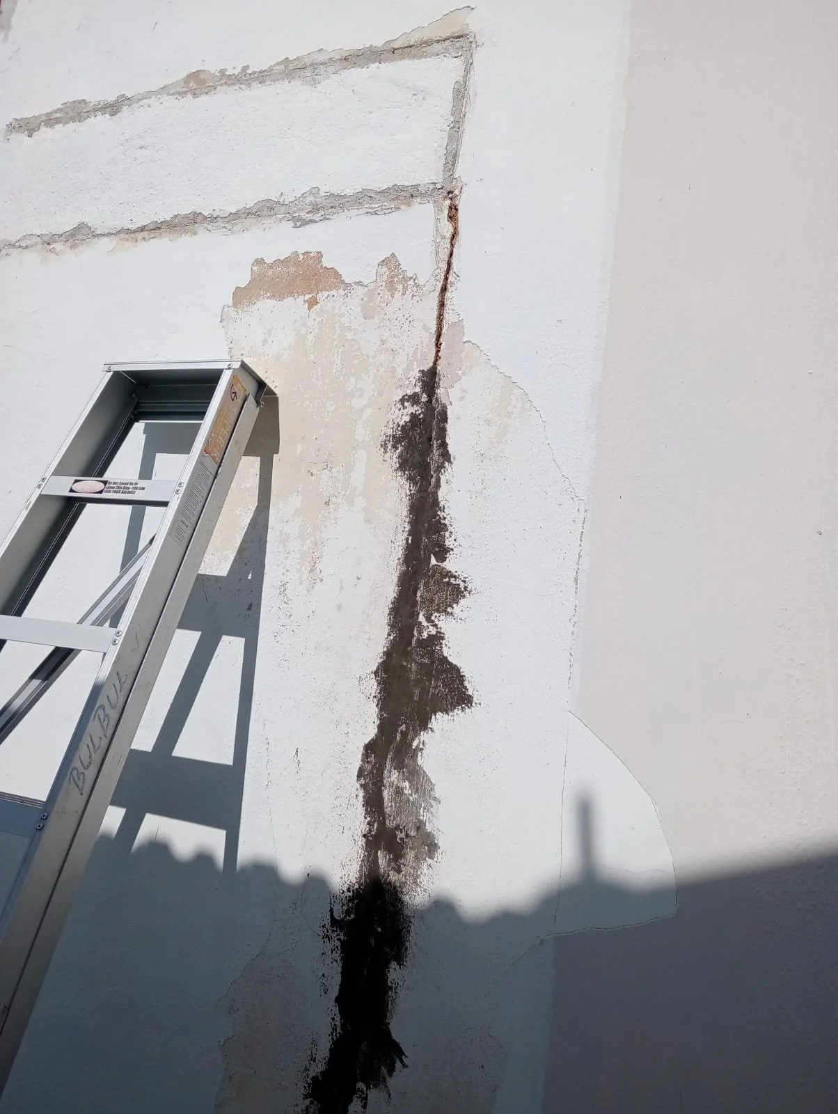 A damaged wall with a vertical crack and peeling paint, showing exposed material beneath. A silver extension ladder leans against the wall.