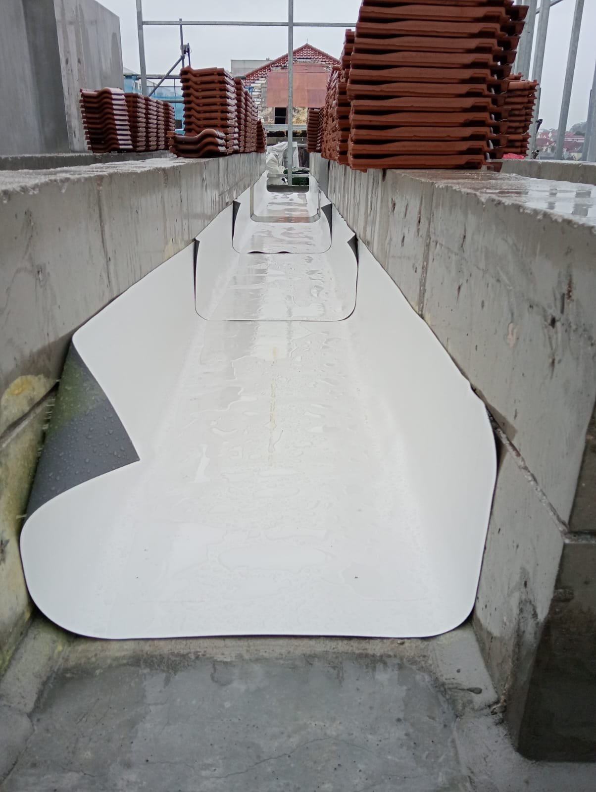 Construction site with sloped concrete wall with a metal sheet lining, stacks of red roof tiles on top, scaffolding in background, and a building with a red-tiled roof in the distance.