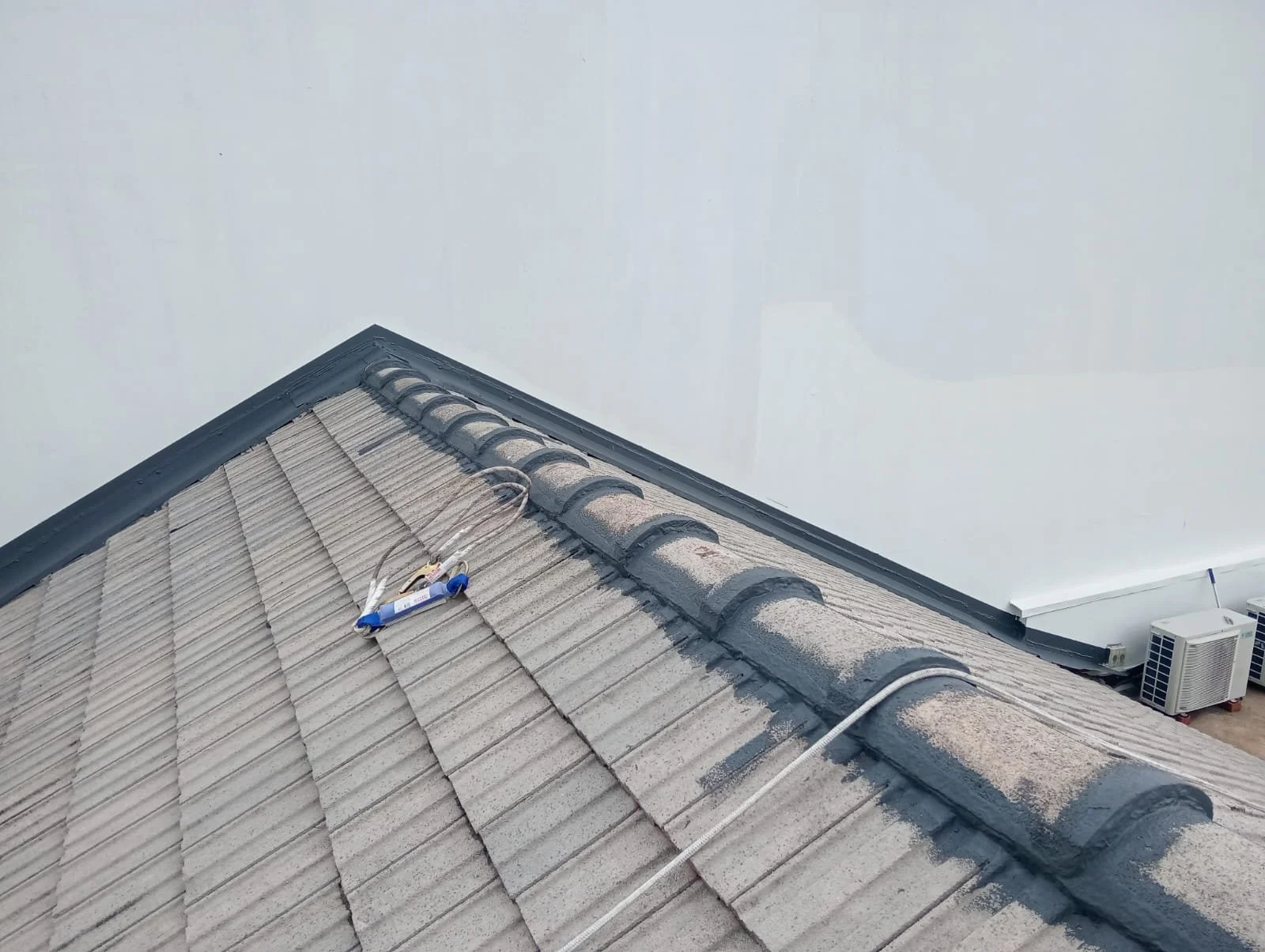 Close-up of a tile roof on a building with a grey vent pipe and electrical wire, with a white wall in the background.
