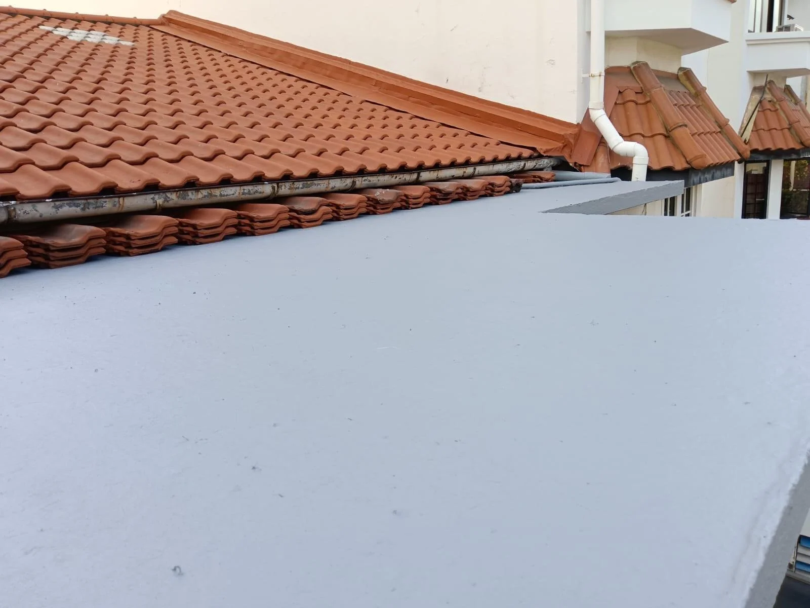 Close-up of a rooftop with orange clay tiles, a section of new white flat roof, and a white gutter pipe against a building with additional rooftop sections in the background.