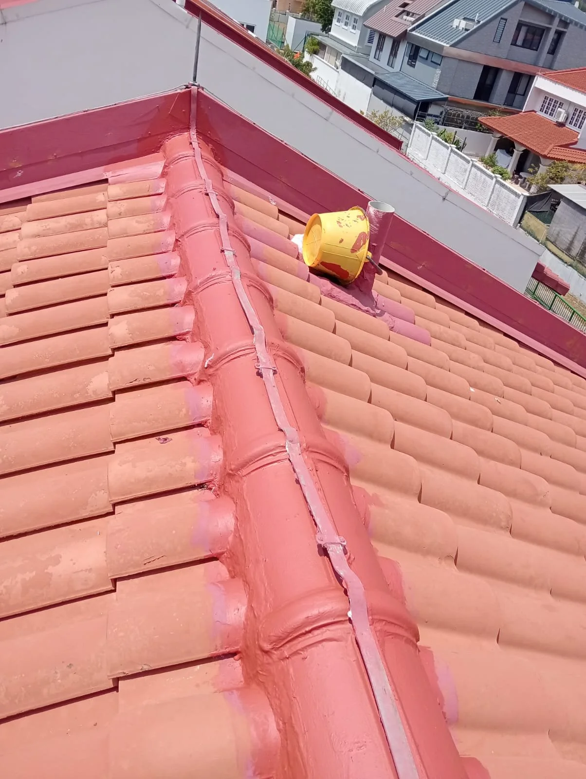 View of a tiled rooftop with a yellow vent pipe, a metal pipe, and pink sealant around the ridges, with neighboring houses in the background.