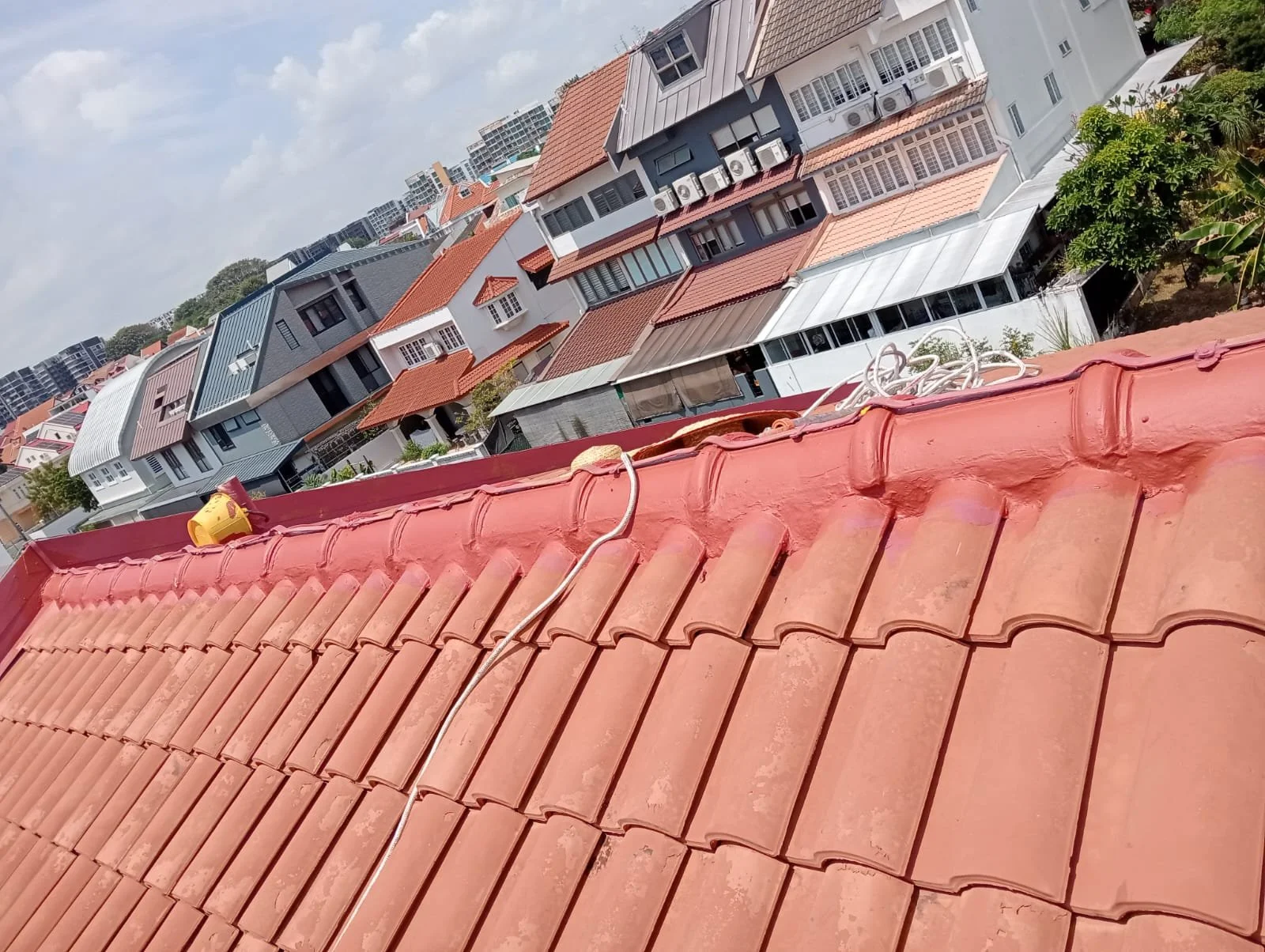 View of rooftops of various houses with red, gray, and white tiles in a neighborhood, taken from a rooftop with a red tile surface in the foreground.