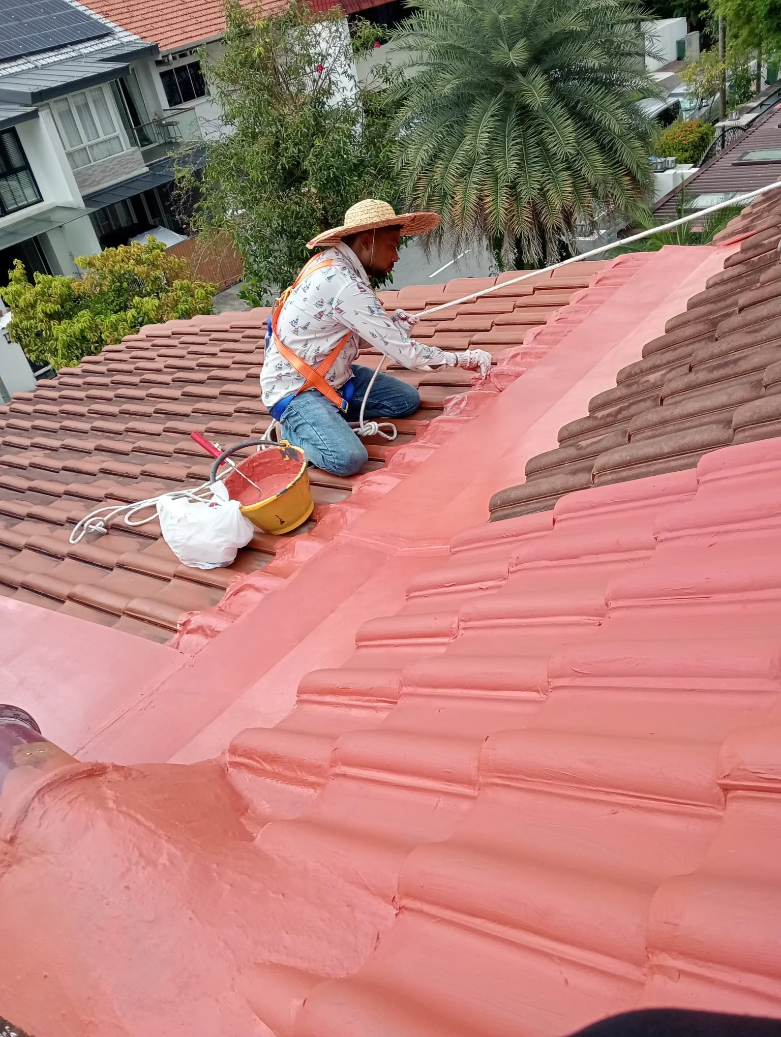A man wearing a large straw hat, safety harness, and gloves, kneeling on a red-tiled roof, painting tiles with a paintbrush. There are trees and residential buildings in the background.