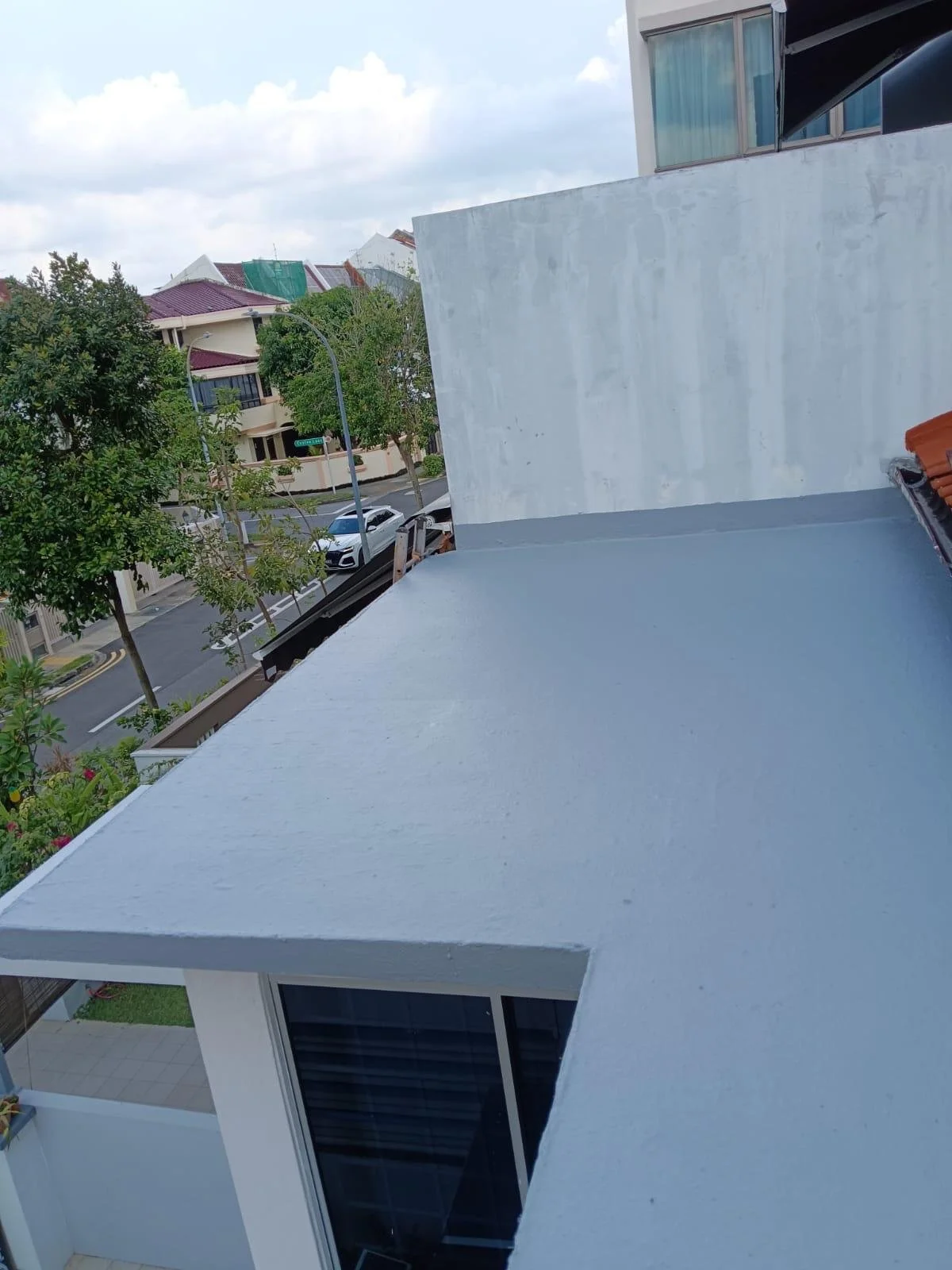 View from a balcony showing a rooftop, trees, parked cars, and neighboring buildings with a cloudy sky.