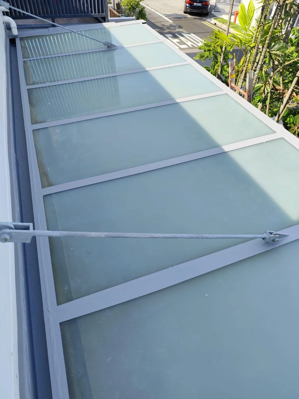 View of a glass roof with white framing, angled upwards, overlooking trees and a street with parked cars.