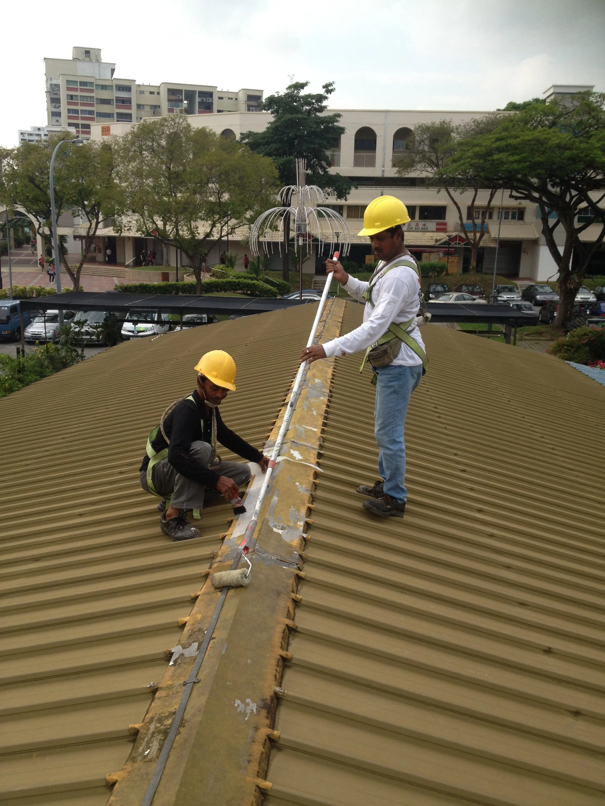 Two construction workers wearing yellow safety helmets working on a metal roof. One worker is kneeling and the other is standing, both handling a long metal pipe. The background shows trees, buildings, and parked cars.