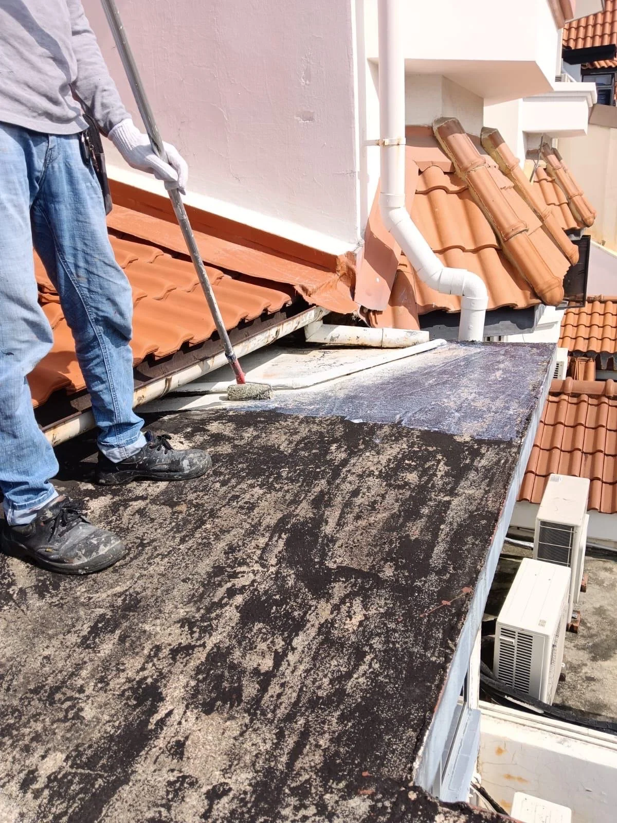 A person cleaning a roof with a roller brush, wearing gloves, jeans, and work boots, with tiles and vents visible.
