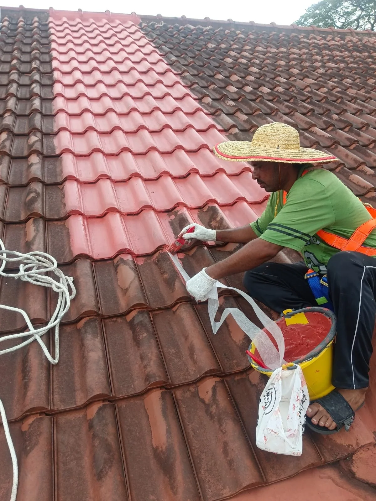 A man wearing a large straw hat, green shirt, and safety harness is painting a section of a red clay tile roof with a brush. A bucket of paint and protective gloves are nearby.