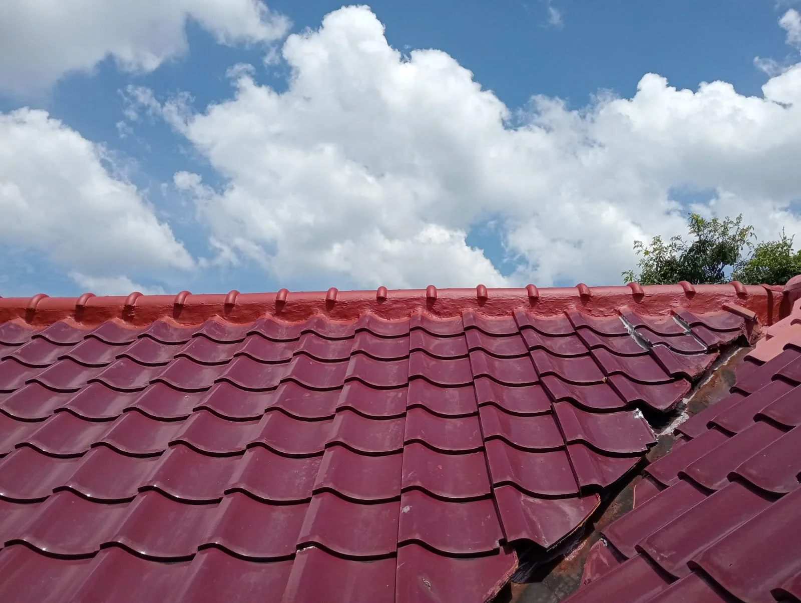 Red roof tiles with damaged tiles in the upper right corner, blue sky with white clouds, and some trees in the background.