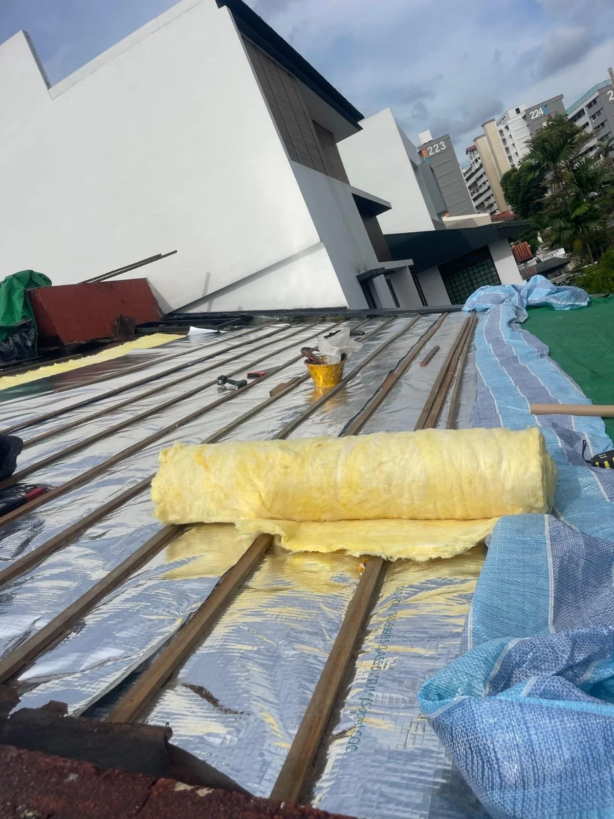 Construction site on a building roof with insulation material, tools, and a tarp, with neighboring buildings visible in the background.