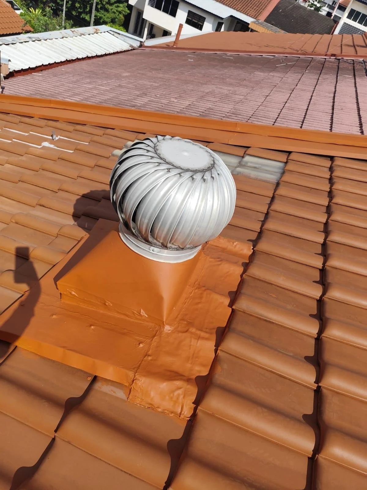 A metal roof vent pipe protrudes from a reddish-brown tiled roof, with neighboring rooftops visible in the background under a clear sky.